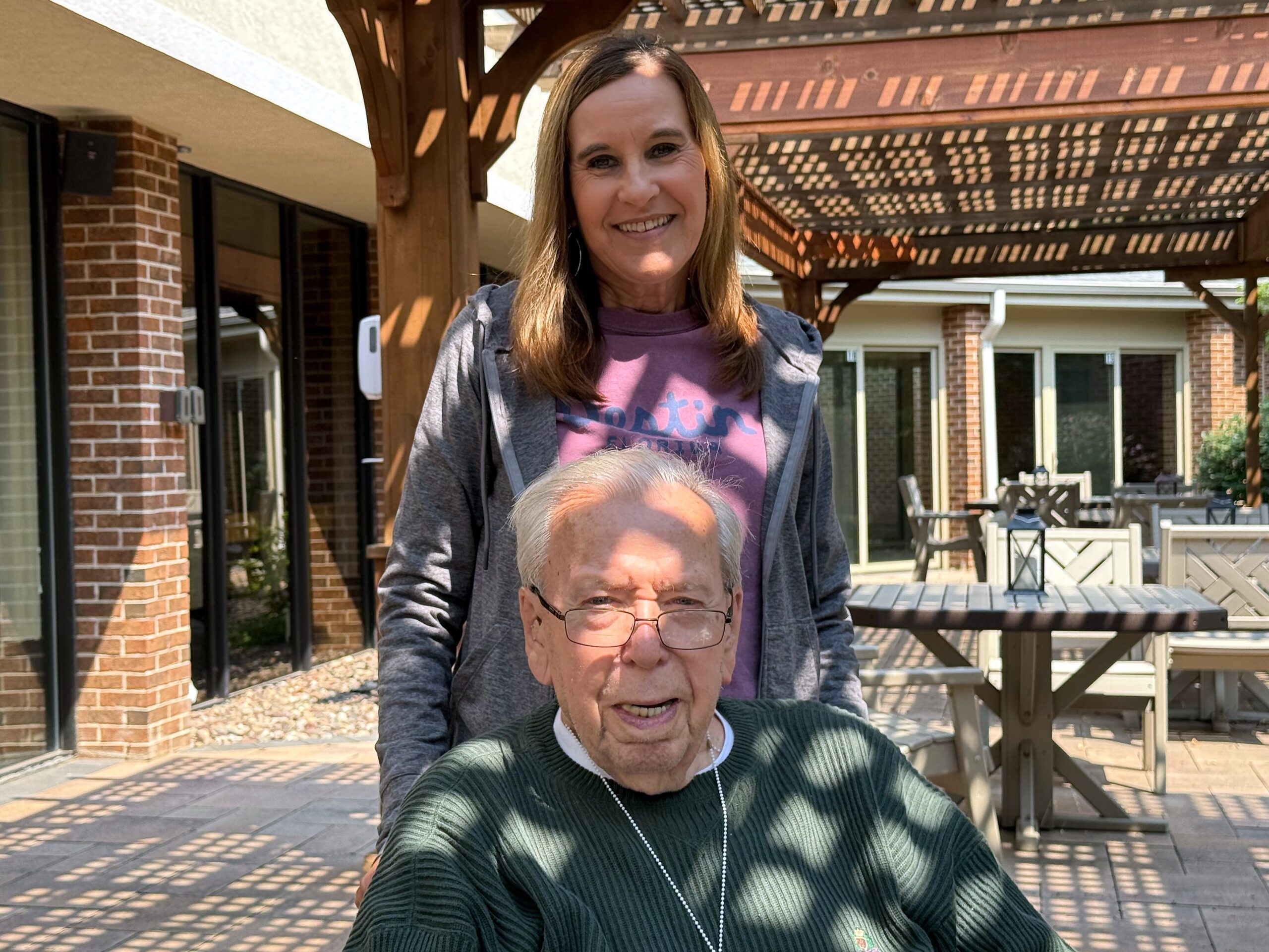 Smiling woman standing behind an elderly man, posing for a selfie on a sunny outdoor patio with brick buildings and a wooden pergola.