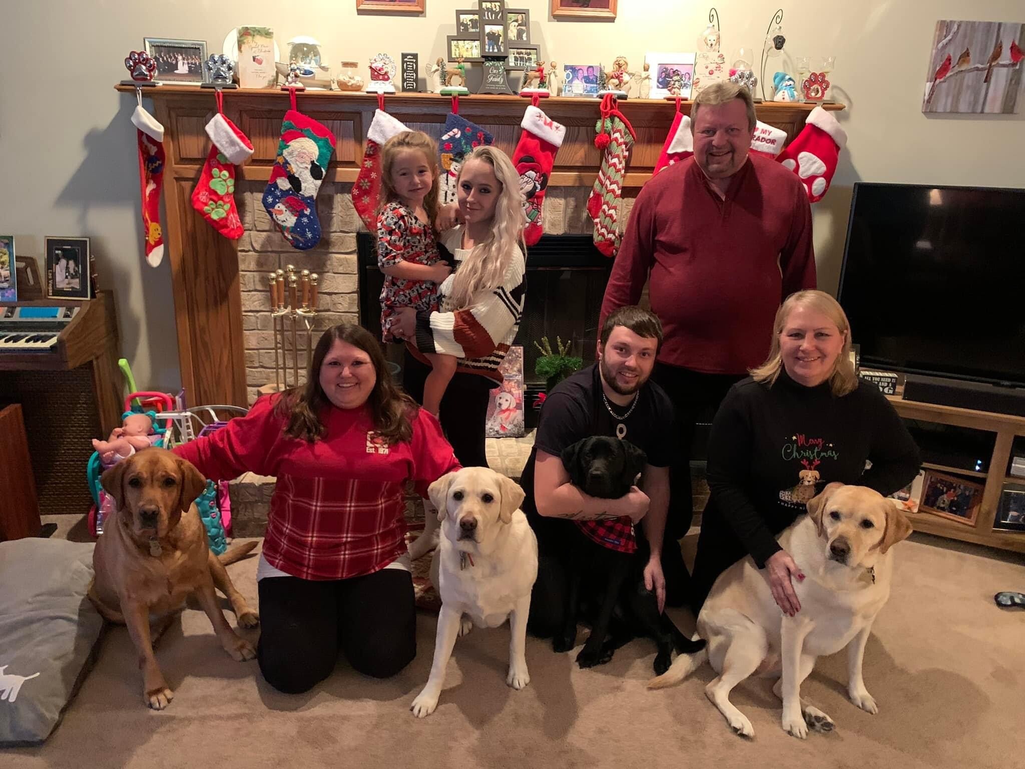 Family group photo in a living room with Christmas stockings, smiling adults and several dogs on the floor in front of a fireplace mantel decorated for the holidays