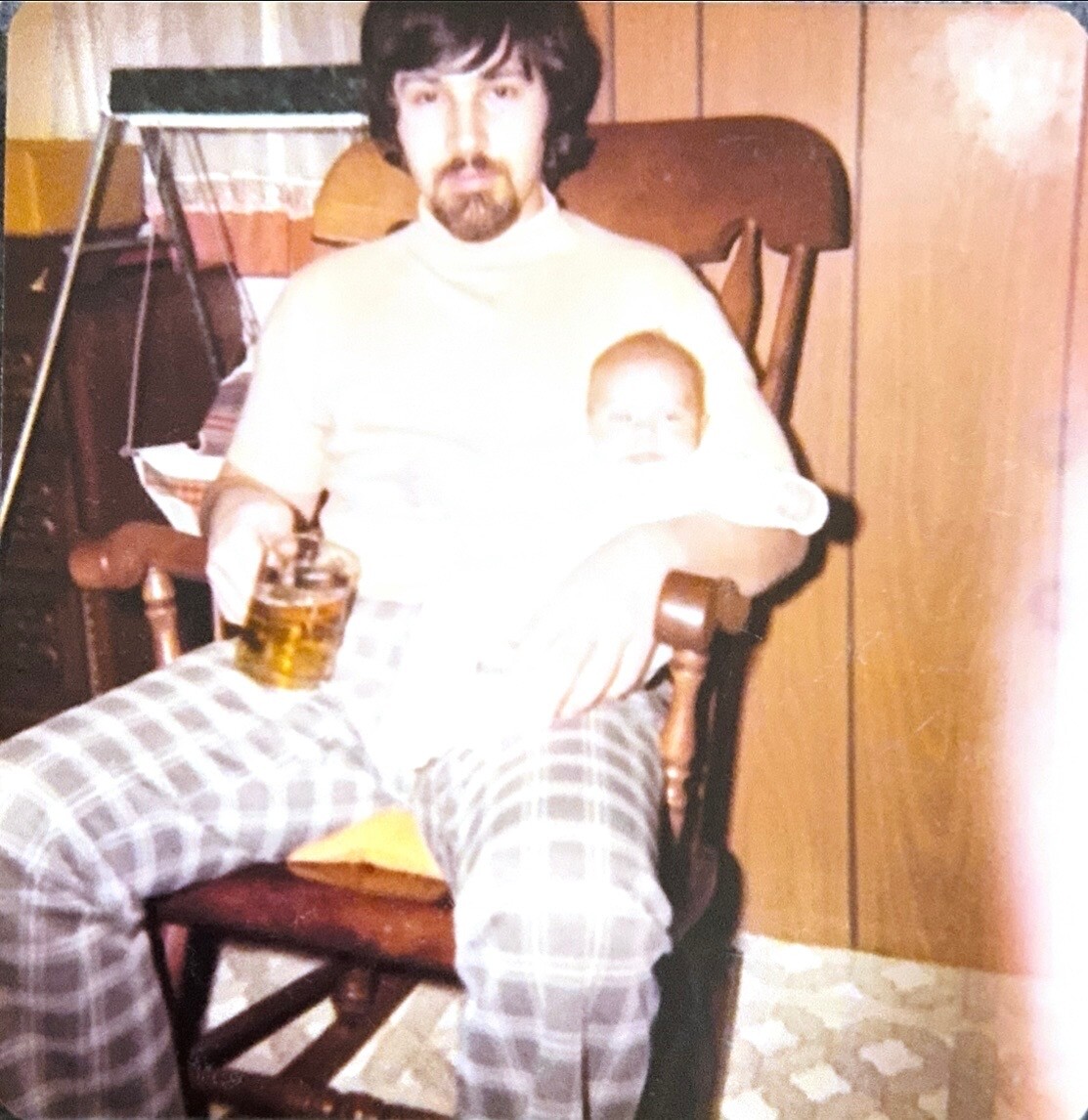 Man with dark hair and beard sitting in a wooden rocking chair, wearing a white turtleneck, holding a baby on his lap and a mug of beer in his hand indoors.