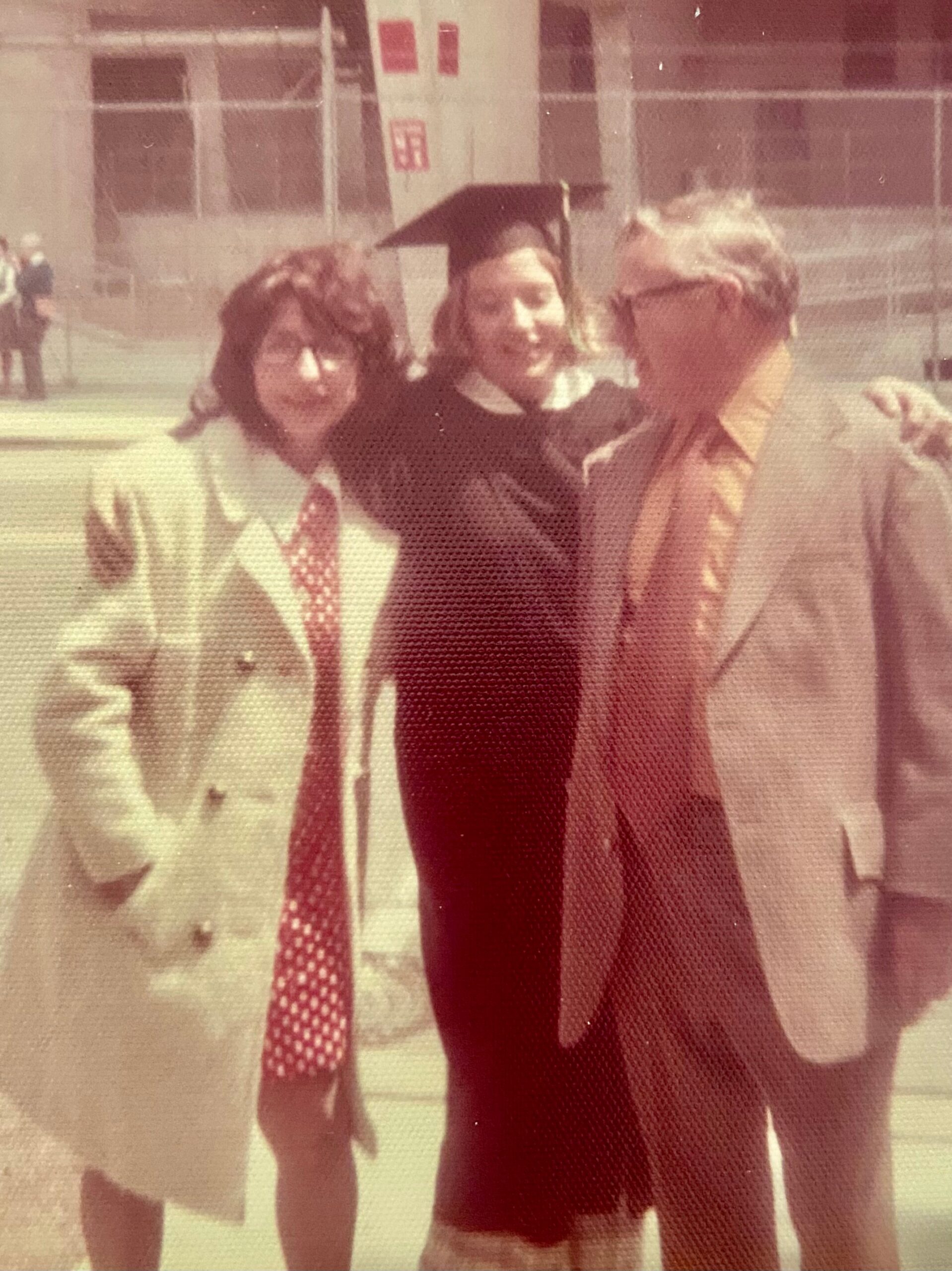 Three people pose outside a building; the central person wears a graduation cap and gown.