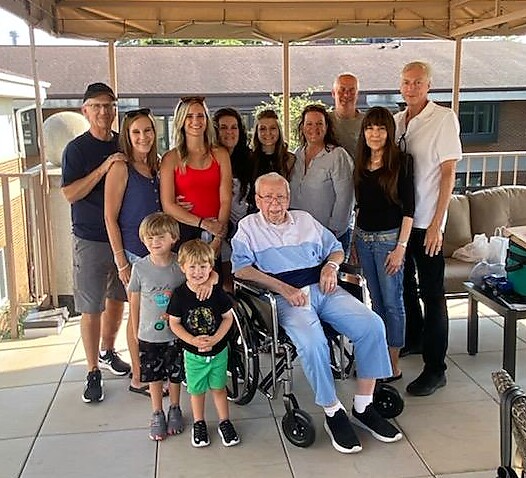 Elderly man in wheelchair at center, surrounded by smiling family on a covered outdoor patio posing for a group photo.