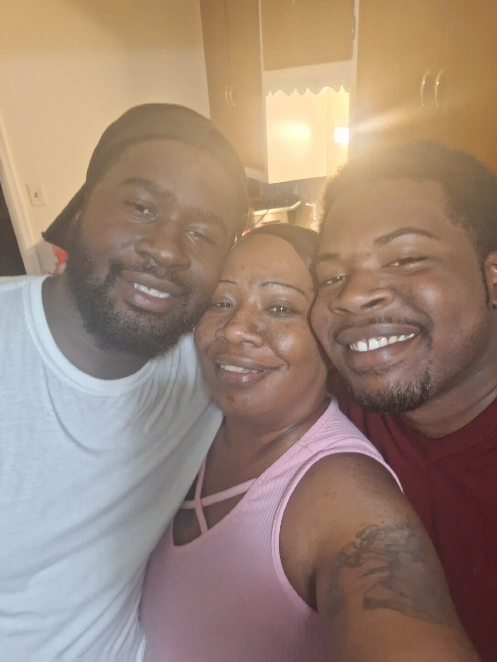 Three adults smiling for a close-up group selfie in a kitchen, with wooden cabinets in the background.