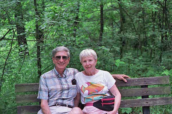 Older couple sitting on a wooden bench in a lush green forest, smiling at the camera.