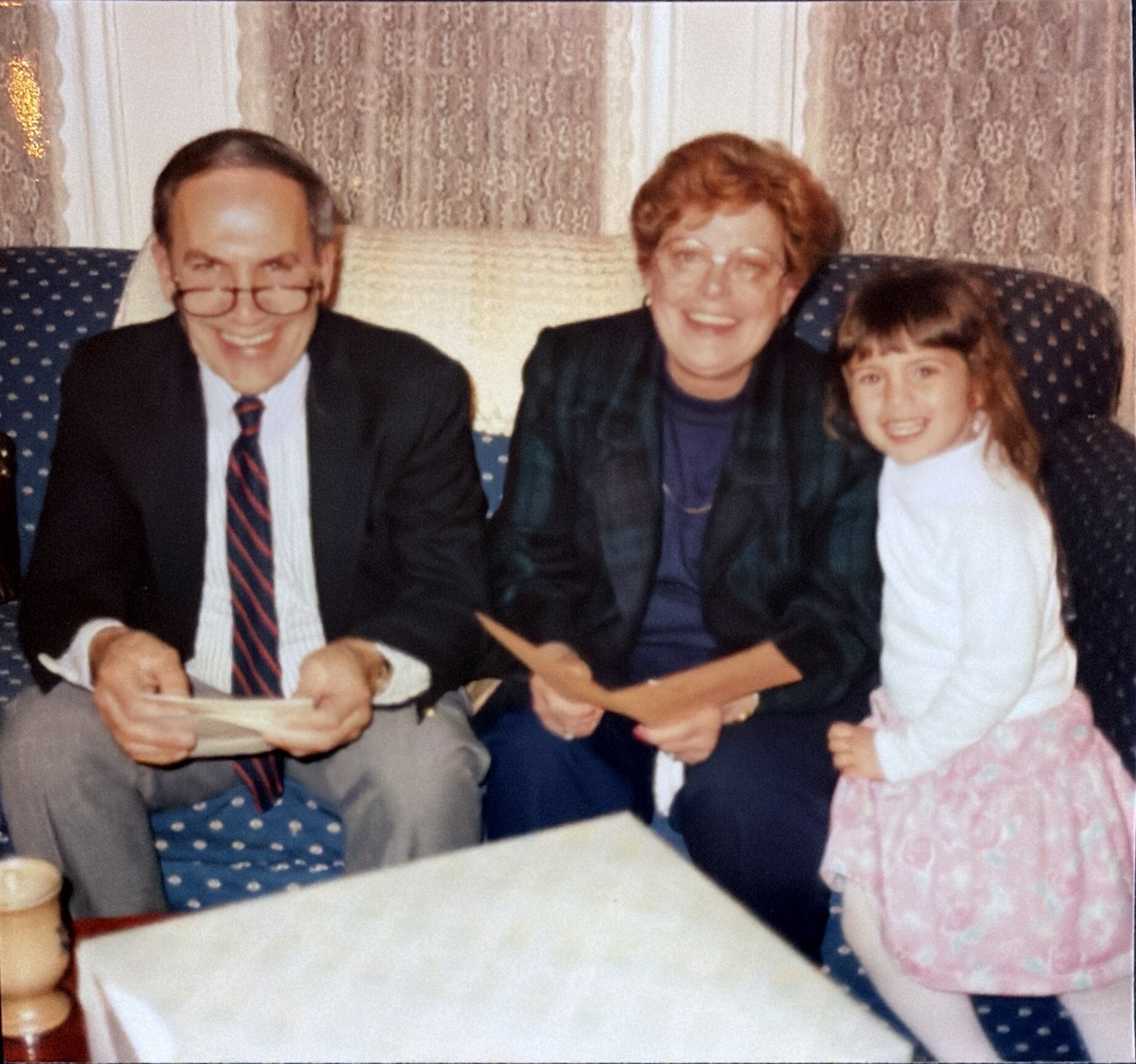 Three people posing on a patterned couch: a man in a suit and glasses, an older woman, and a smiling young girl in a pink dress with white tights.