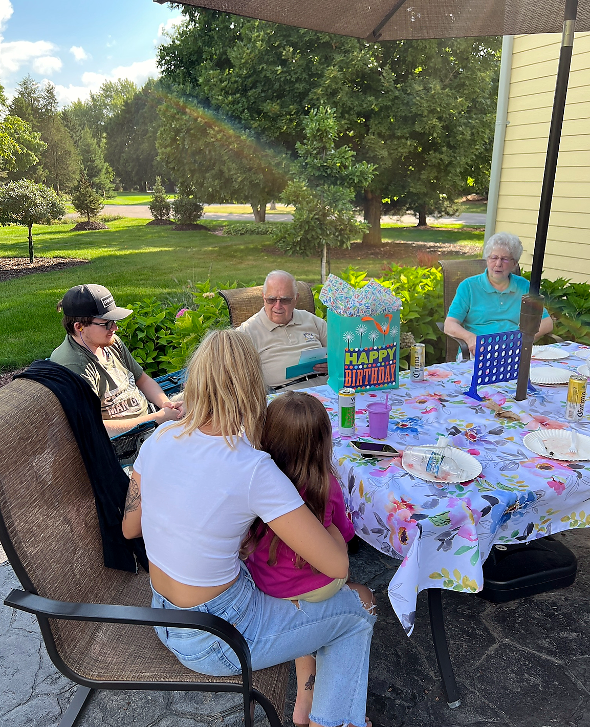 Outdoor birthday party on a patio with a cheery 'Happy Birthday' gift bag and guests seated around a floral tablecloth.