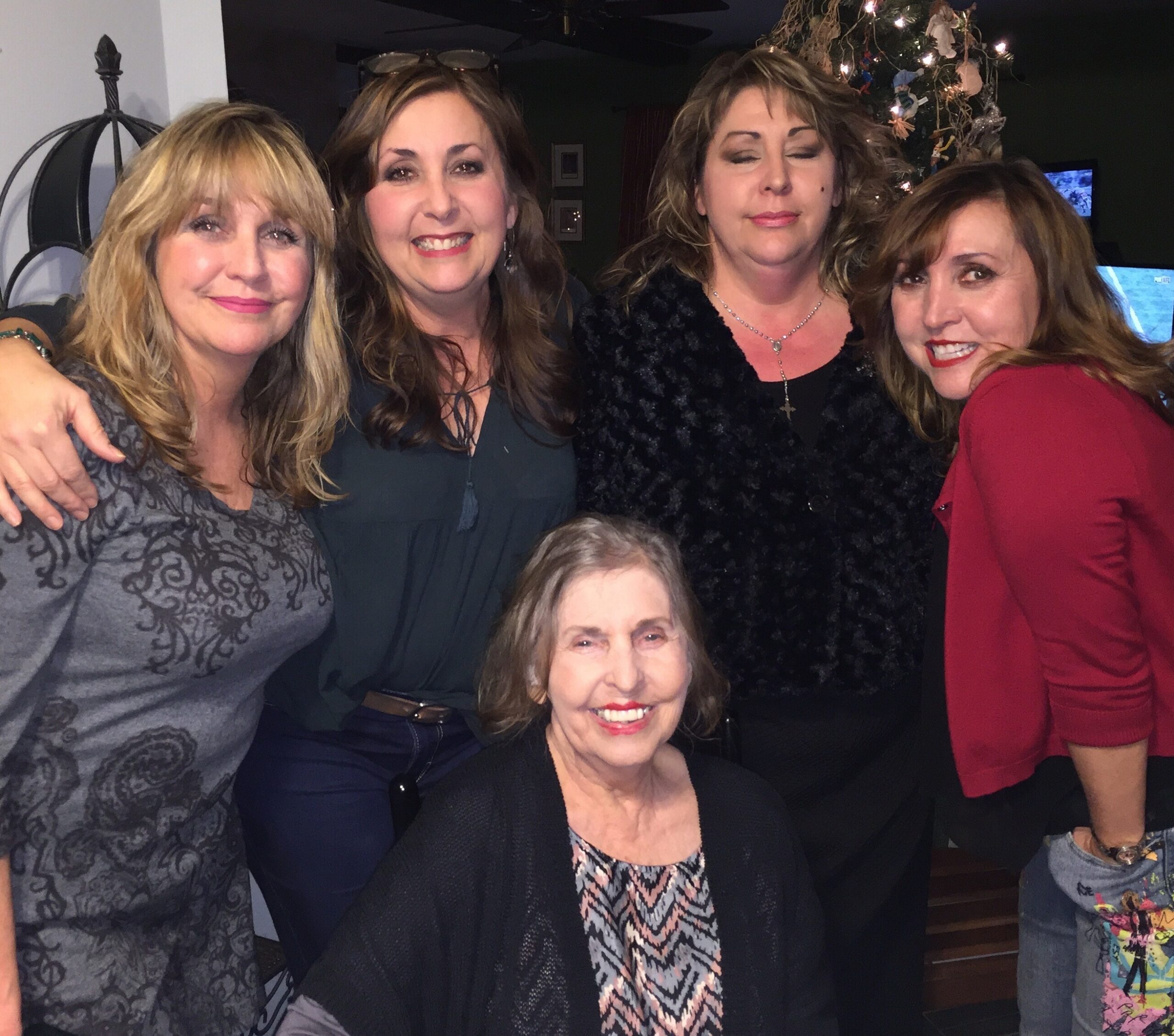 Four women posing indoors at a holiday gathering, smiling, with a decorated Christmas tree in the background.