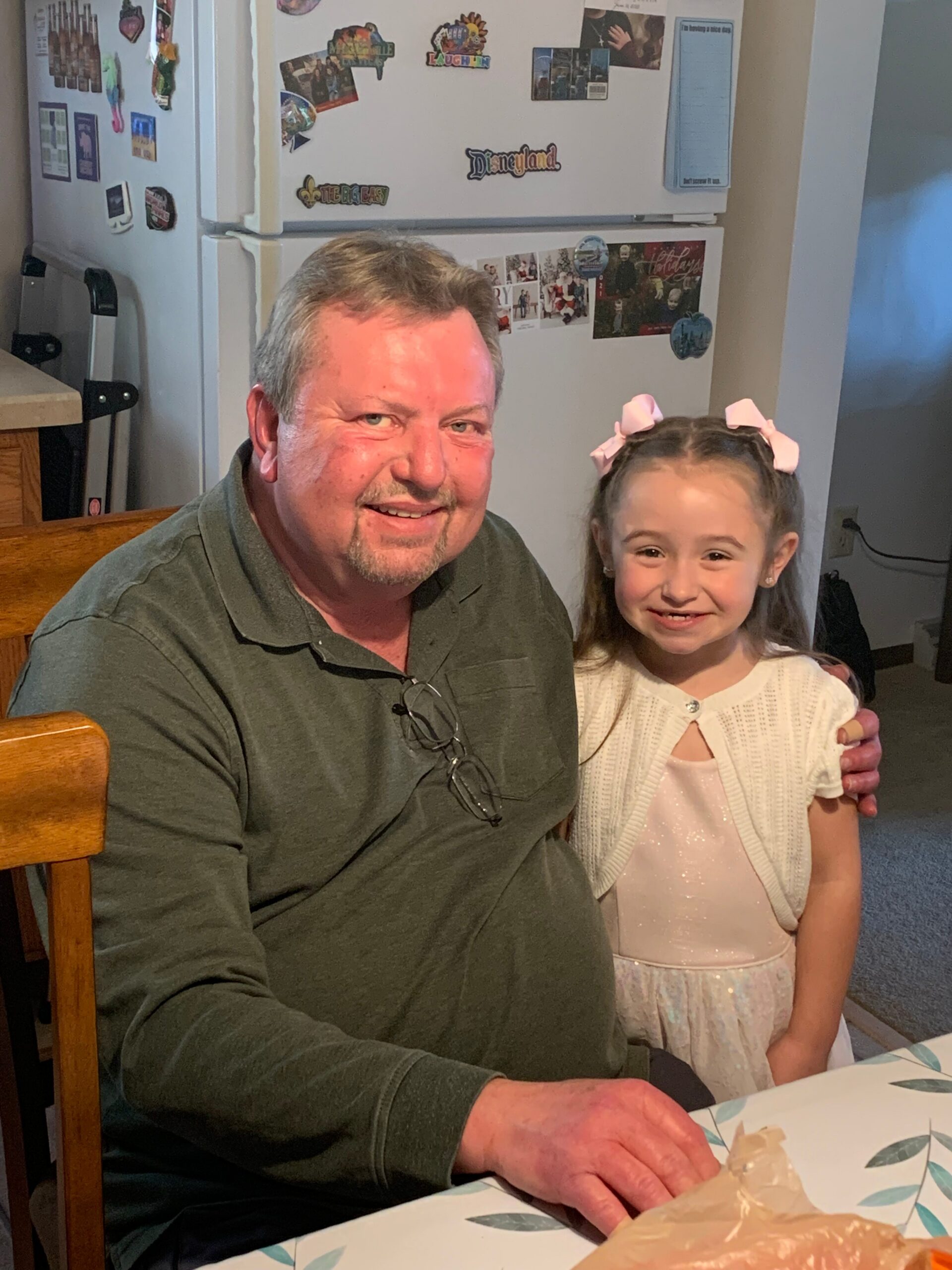 Smiling man and young girl sit side by side at a table in a kitchen, a cake in front of them on the tablecloth behind them a refrigerator with magnets is visible.