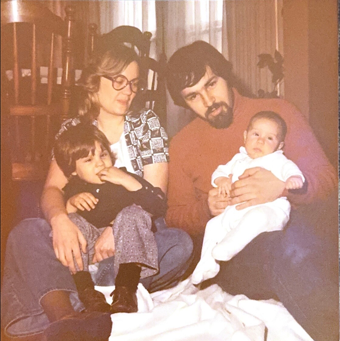 Family portrait: a mom with glasses and a bearded dad sit on a couch with two young children, a toddler on the mom's lap and a baby in the dad's arms, in a cozy home setting.