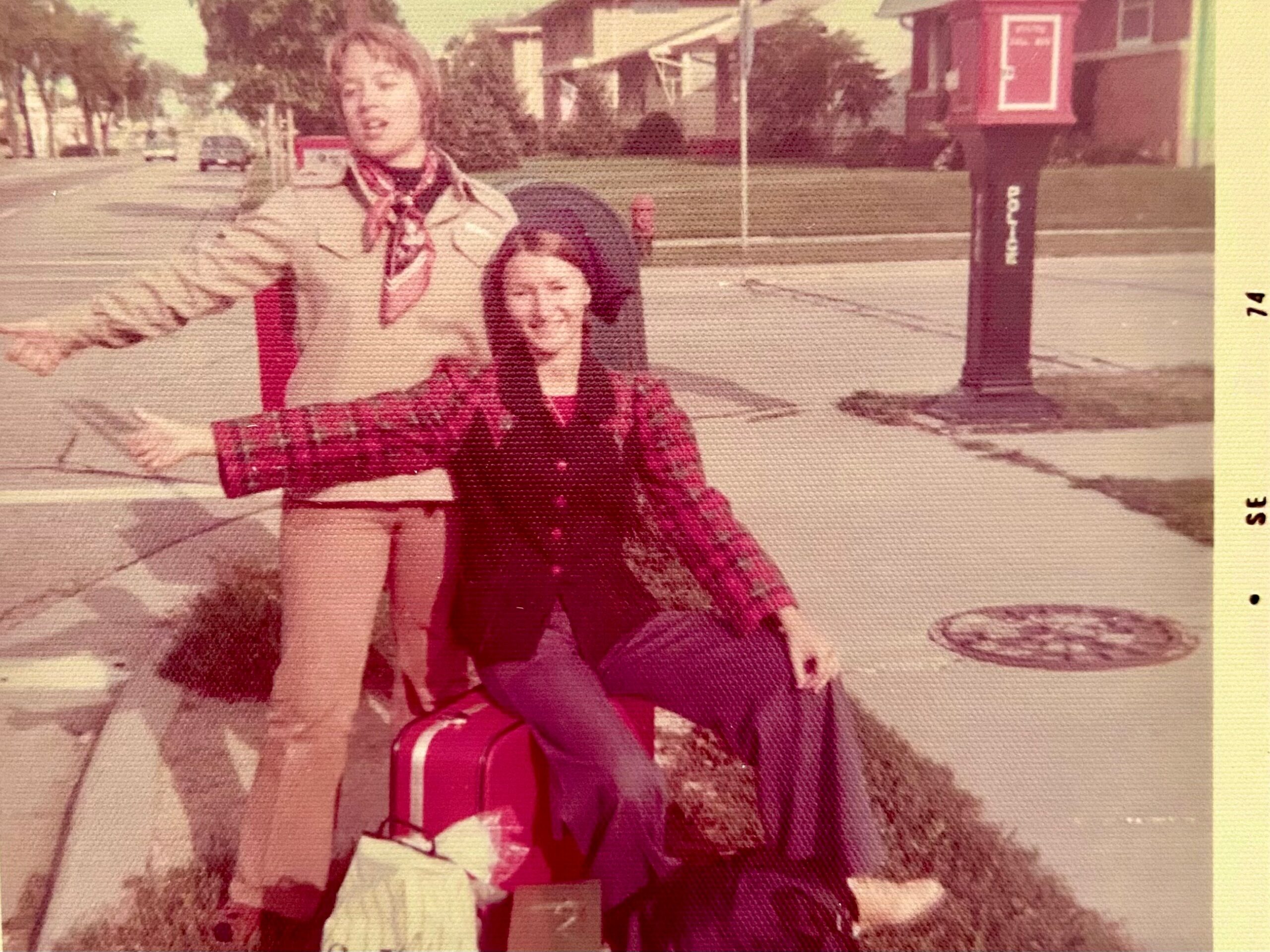 Two kids posing on a residential sidewalk with a street and houses in the background; one in a beige coat and scarf stands behind the other in a red plaid outfit.