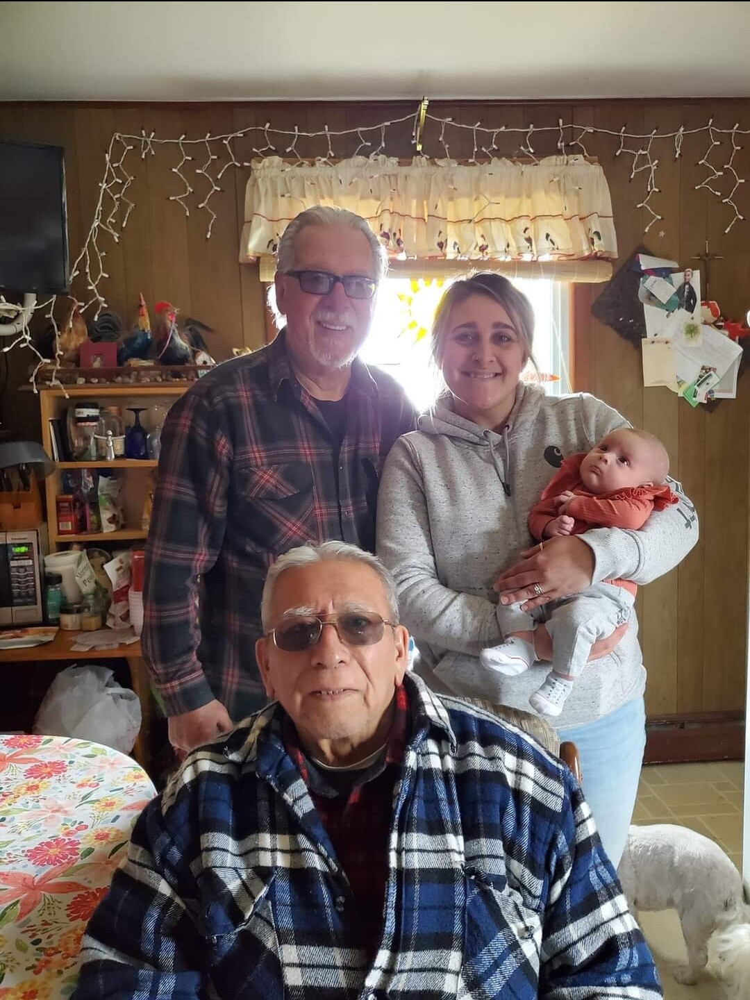 Family group portrait in a cozy kitchen: grandfather in a plaid shirt sits at the table while parents cradle their baby by the window behind him.