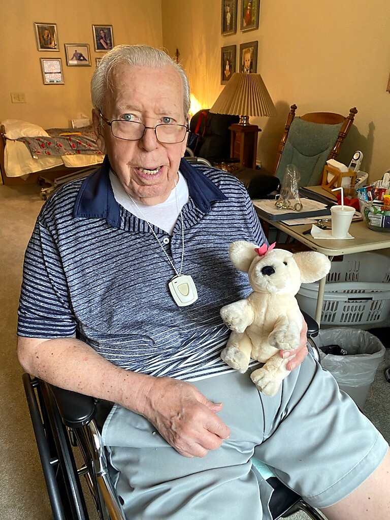 Elderly man in a wheelchair smiling, wearing glasses, holding a small beige teddy bear with a pink bow.