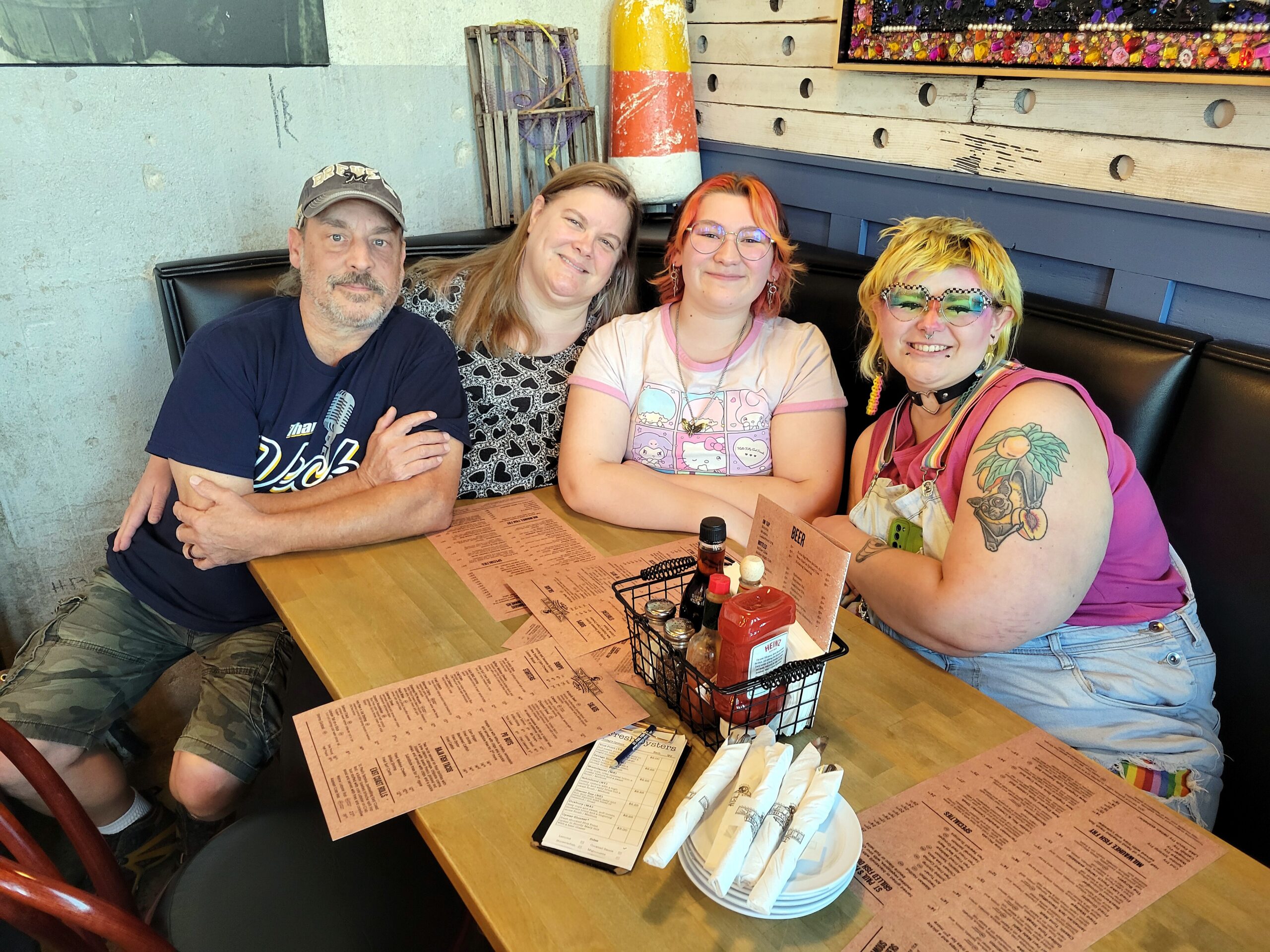 Four friends sit at a booth in a casual restaurant, smiling at the camera with pink menus and condiments on the table in front of them.