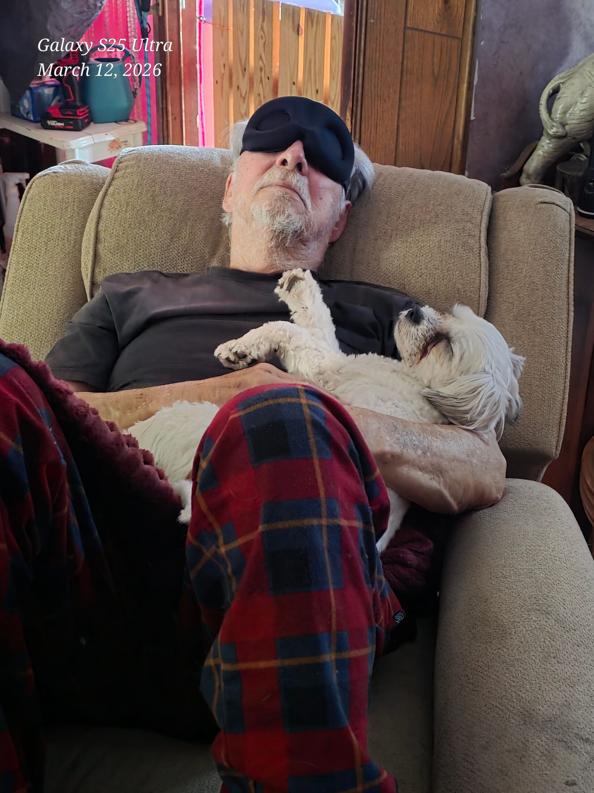 Older man with a white beard sleeping on a beige couch, wearing a dark sleep mask, with a small white dog resting on his chest nearby in a cozy living room.