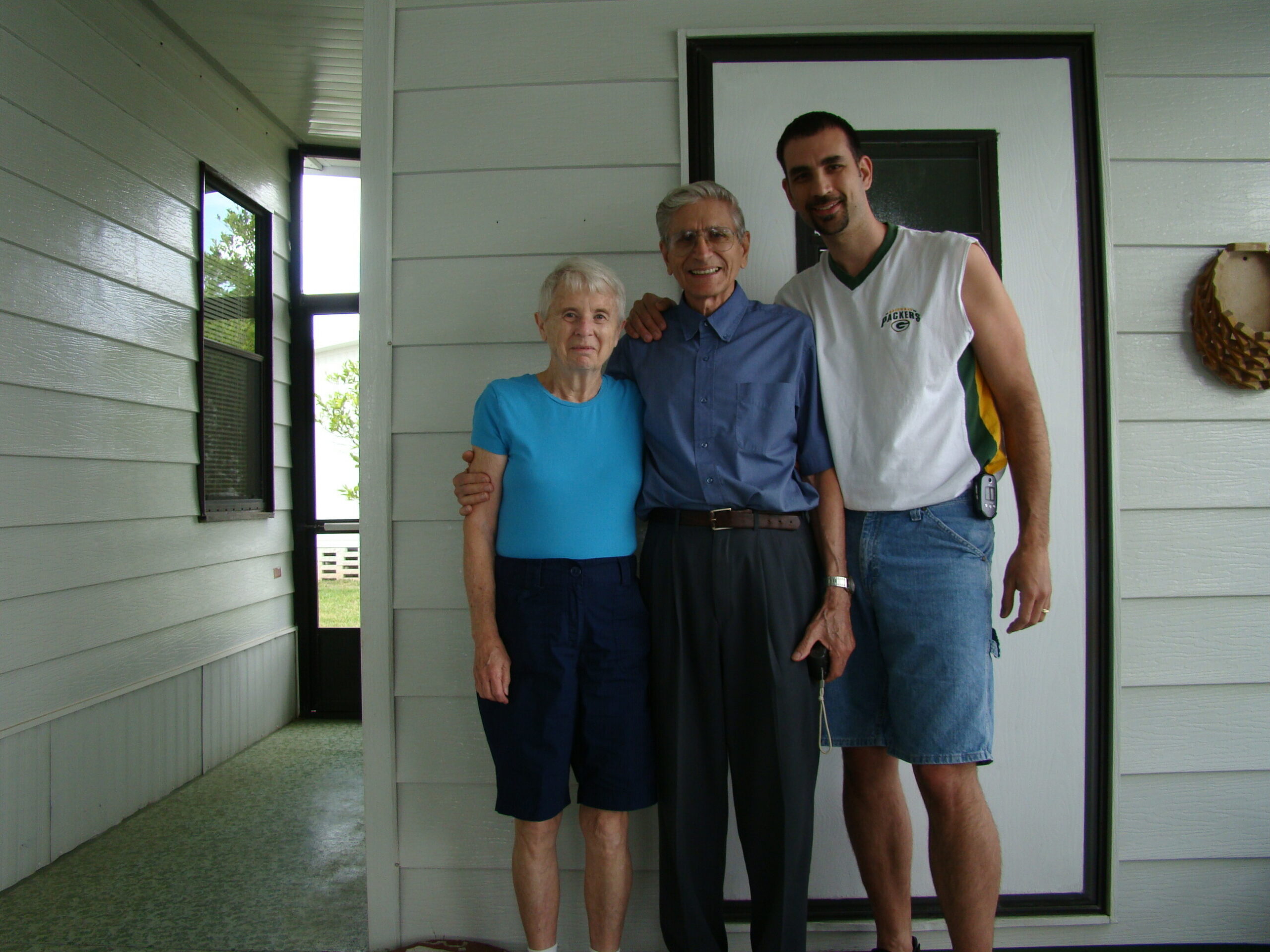 Three adults posing on a front porch: an elderly woman in blue, an elderly man in a blue shirt, and a younger man in a Packers jersey.