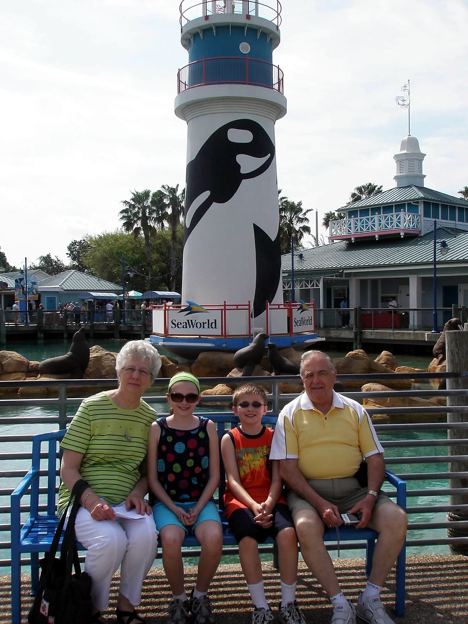 Family of four posing on a blue bench in front of a tall lighthouse-style tower with a large penguin mural at SeaWorld.