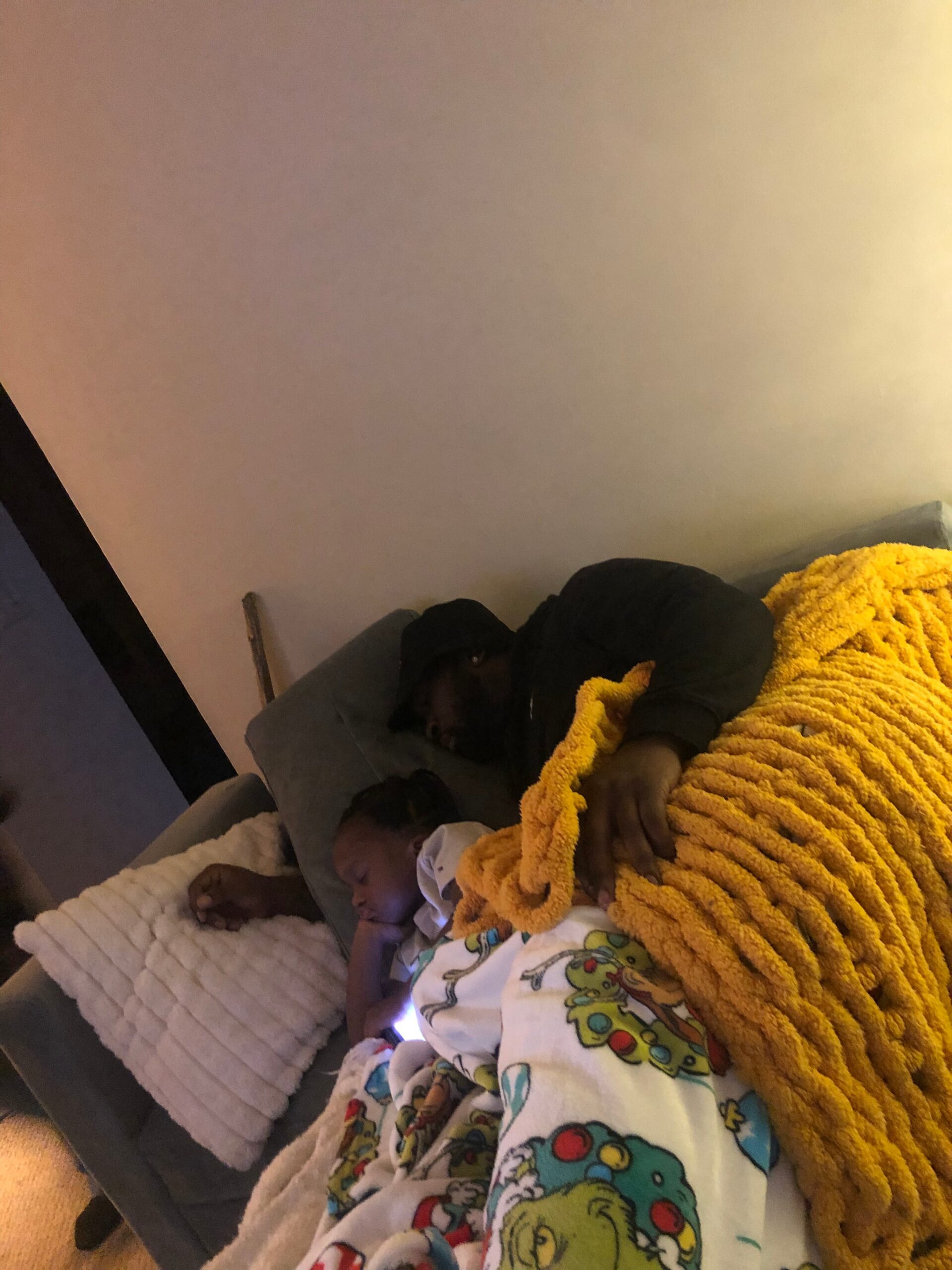 Two children sleeping on a gray sofa, covered with a bright yellow blanket and soft pillows nearby.