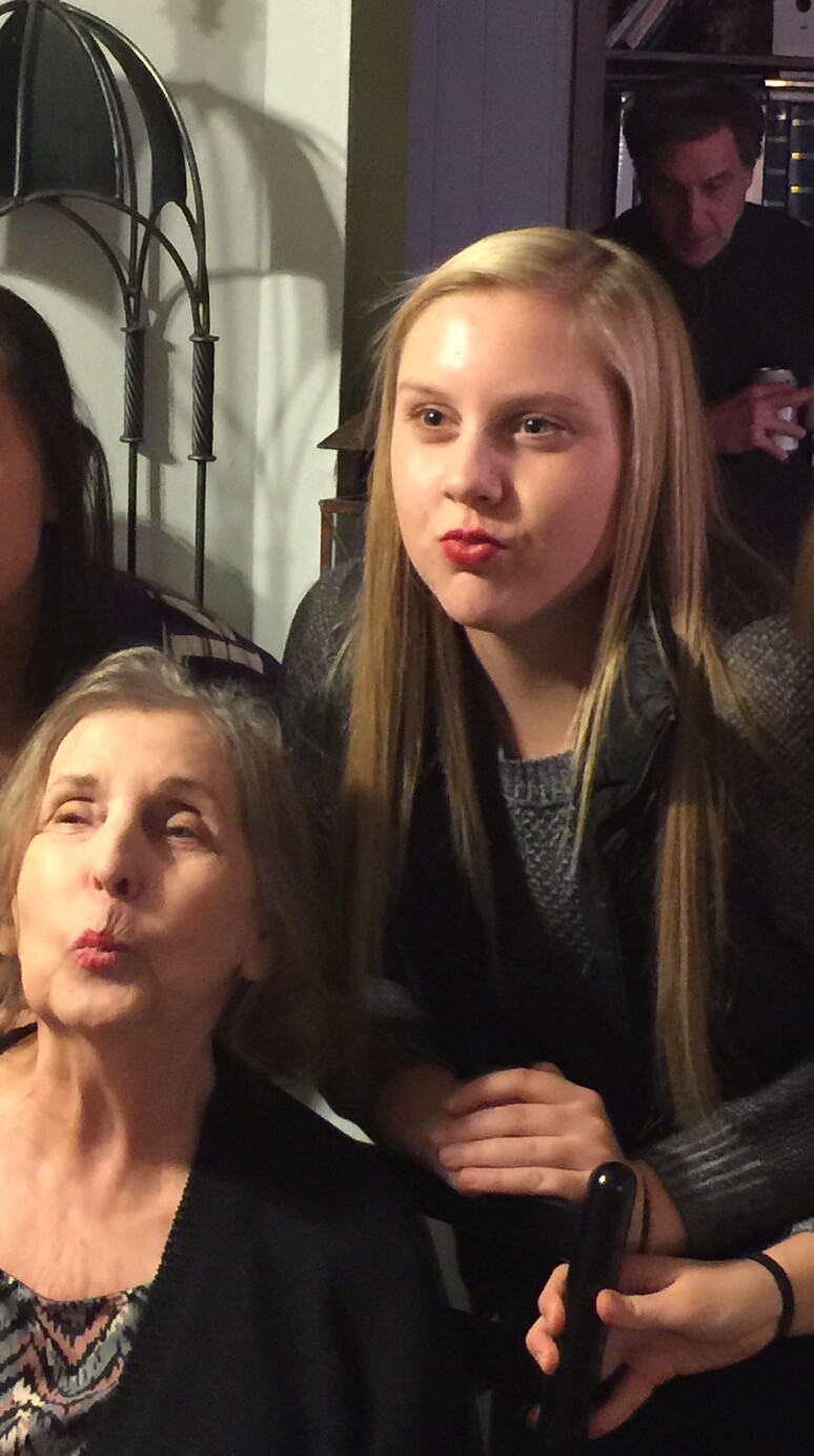 Two women pose for a playful selfie, both puckering their lips toward the camera in a cozy indoor setting, with a man in the background.