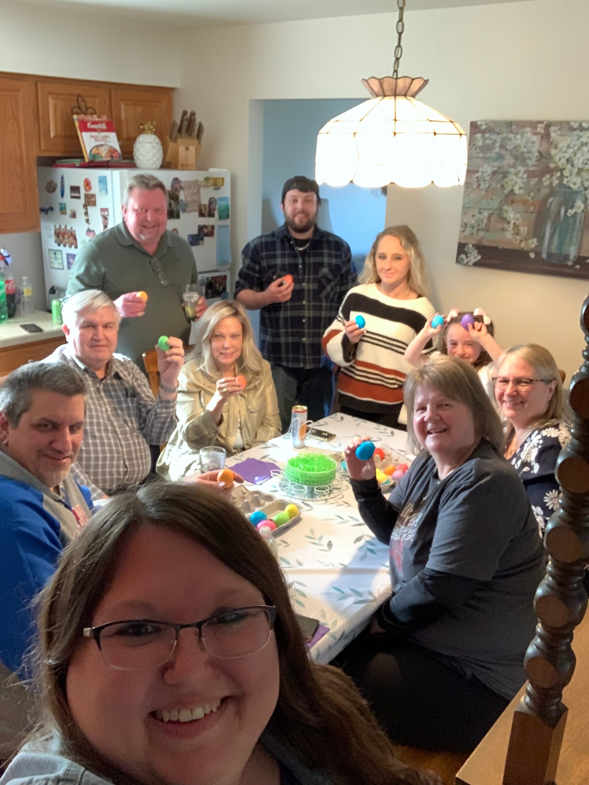 Smiling group around a kitchen table, holding bright dyed Easter eggs as they pose for a photo.