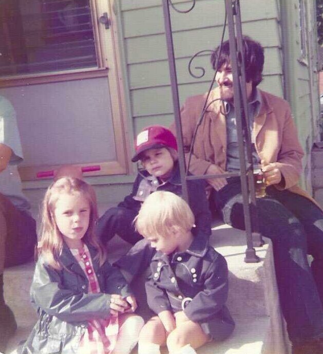 Man with dark hair sitting on a front porch step behind a metal railing, holding a guitar, as three children (two girls and a boy) sit on the steps in front of him.