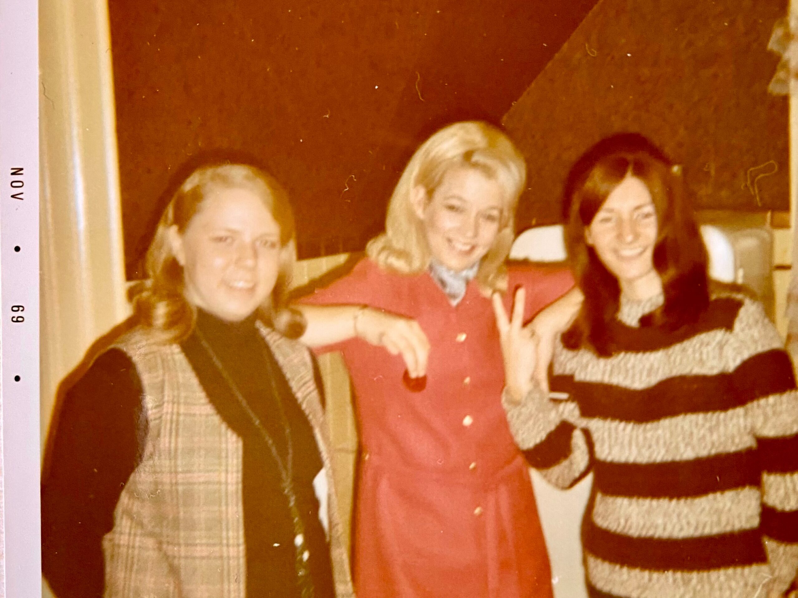 Three women posing together for a photo, smiling, one flashing a peace sign in a warm-toned image (vintage style).