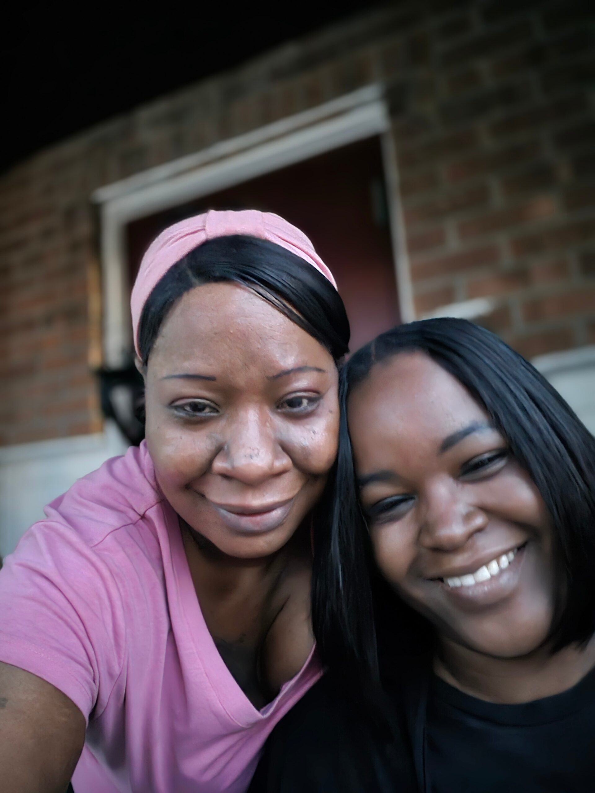 Two women posing for a close-up selfie, smiling in front of a brick wall; one wears a pink headband and pink shirt while the other has long black hair and a black top.