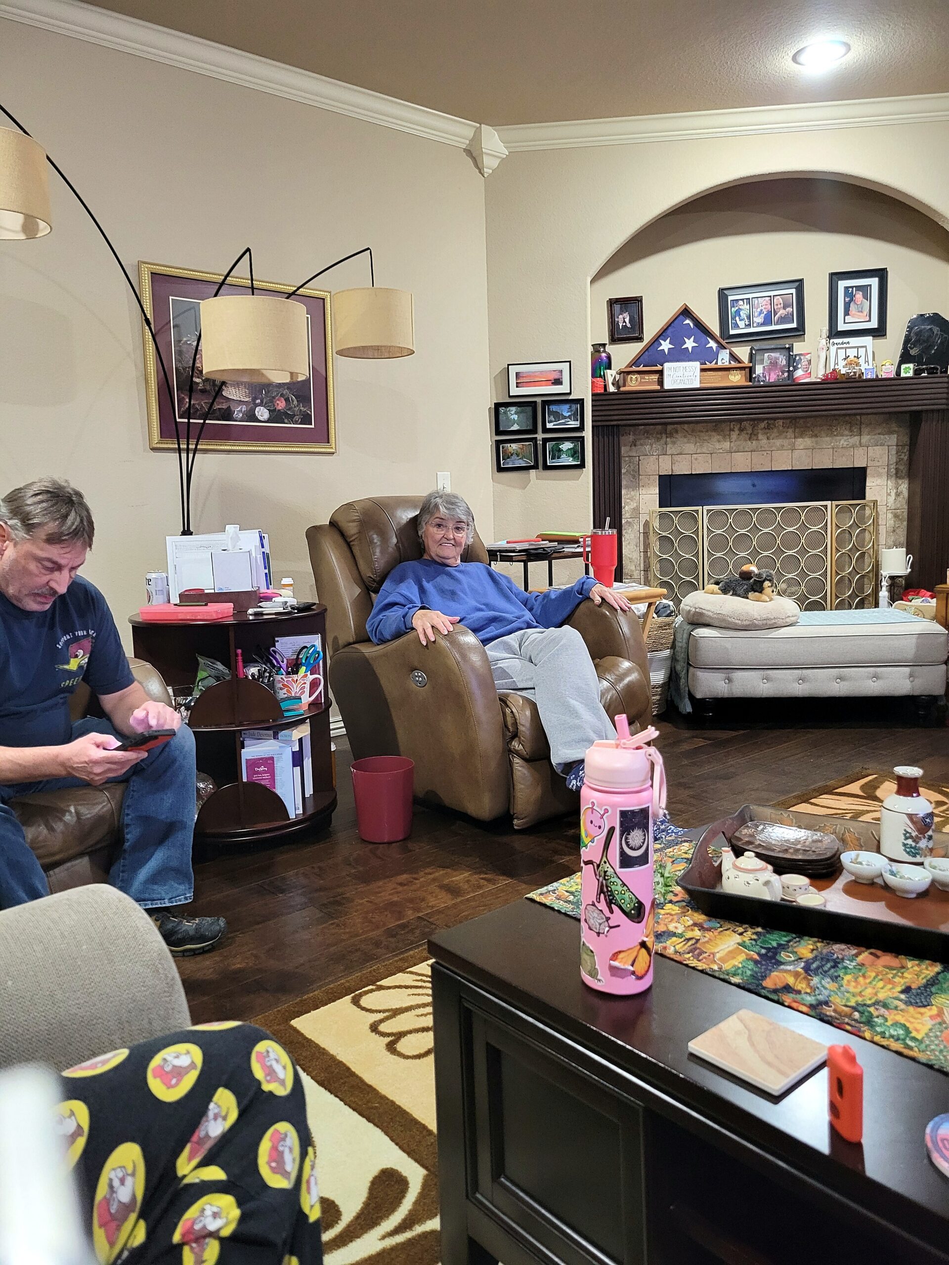 Older woman in a brown recliner wearing blue sits smiling in a cozy living room with a fireplace and wall photos behind her.