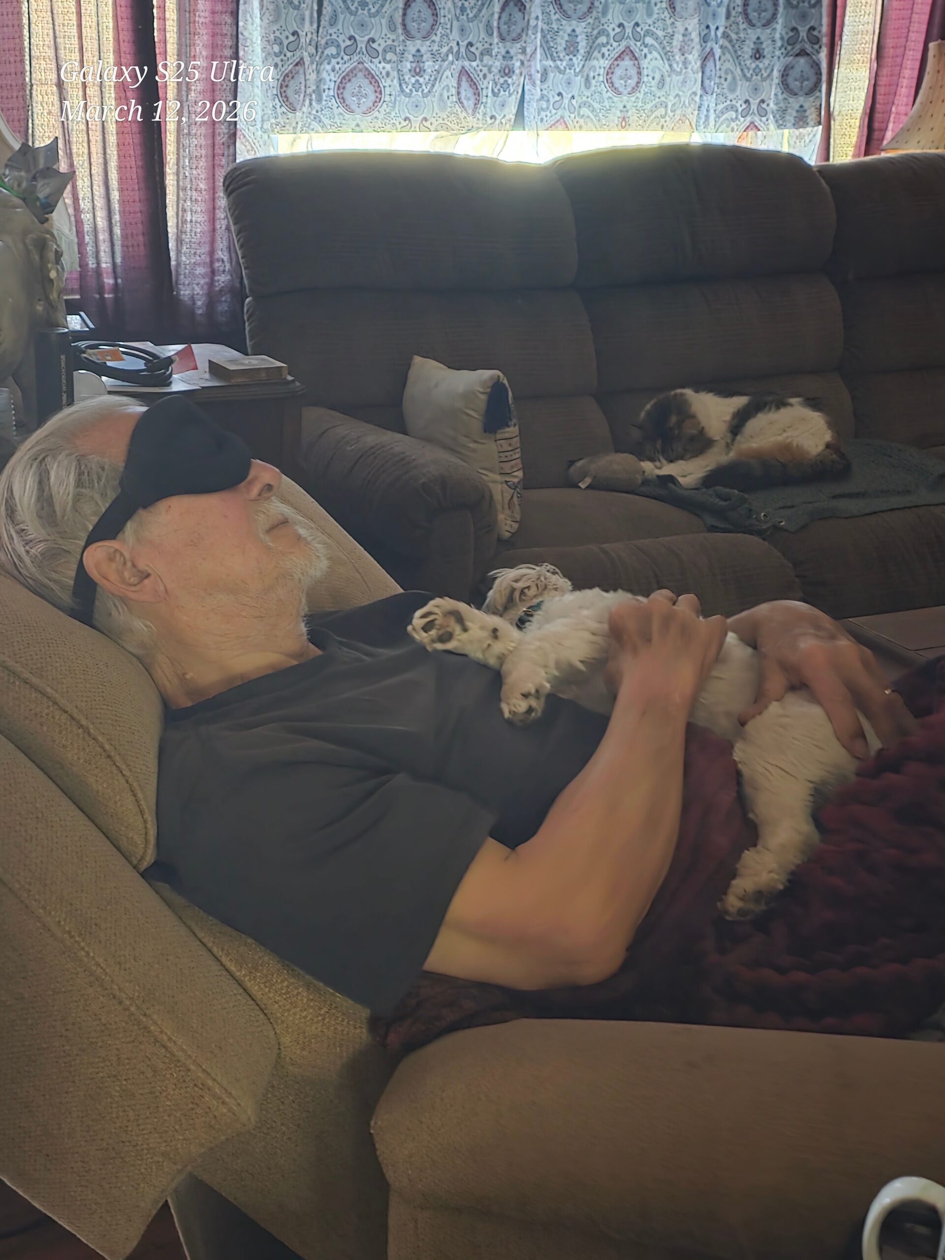 Older man with an eye mask asleep on a brown couch, a small fluffy dog resting on his chest being held.