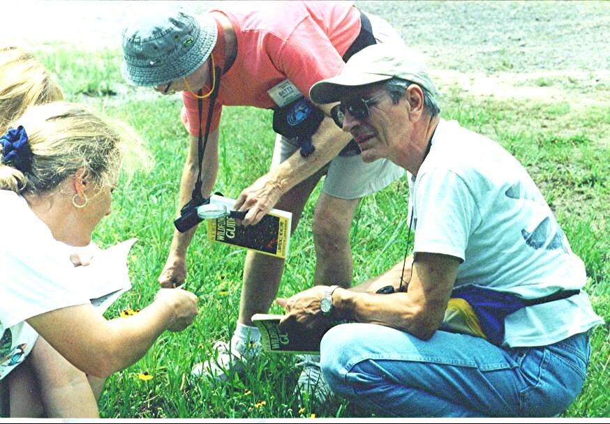 Three people crouch in a grassy field, looking through field guides and a notebook together.