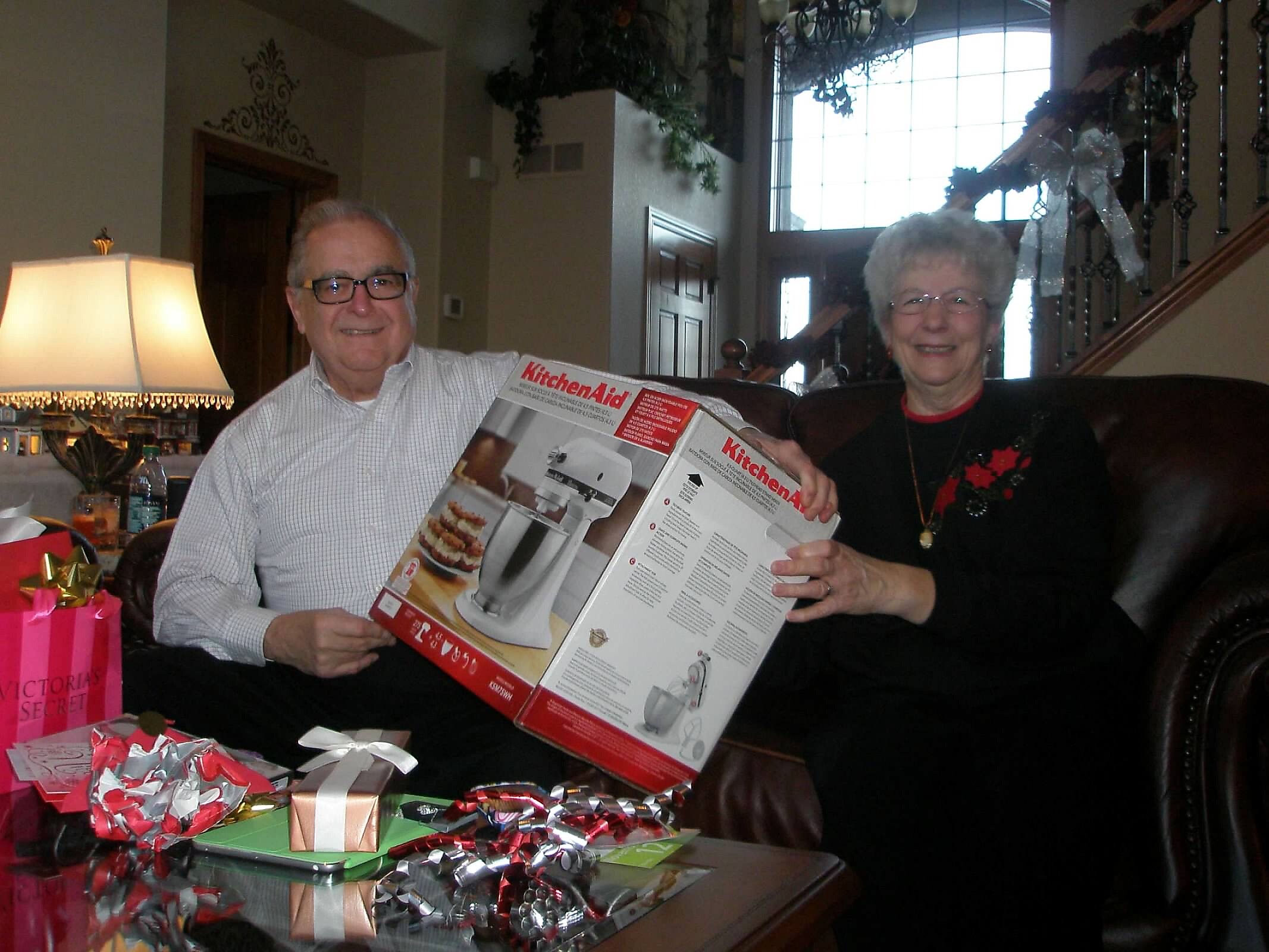 Older couple sitting on a couch, holding a KitchenAid mixer box, surrounded by gift boxes at home Christmas setting.