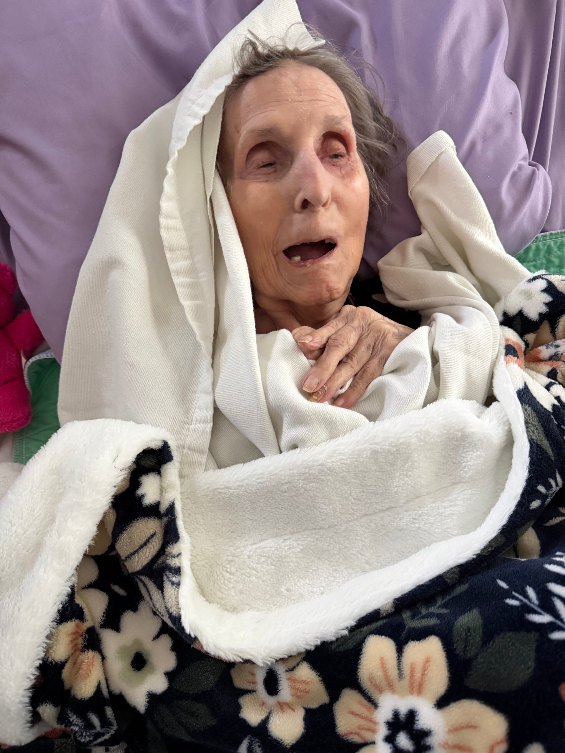 Elderly person wrapped in a white blanket, lying in bed with a purple sheet behind, mouth slightly open and eyes half-closed, resting or unwell in a close-up pose.