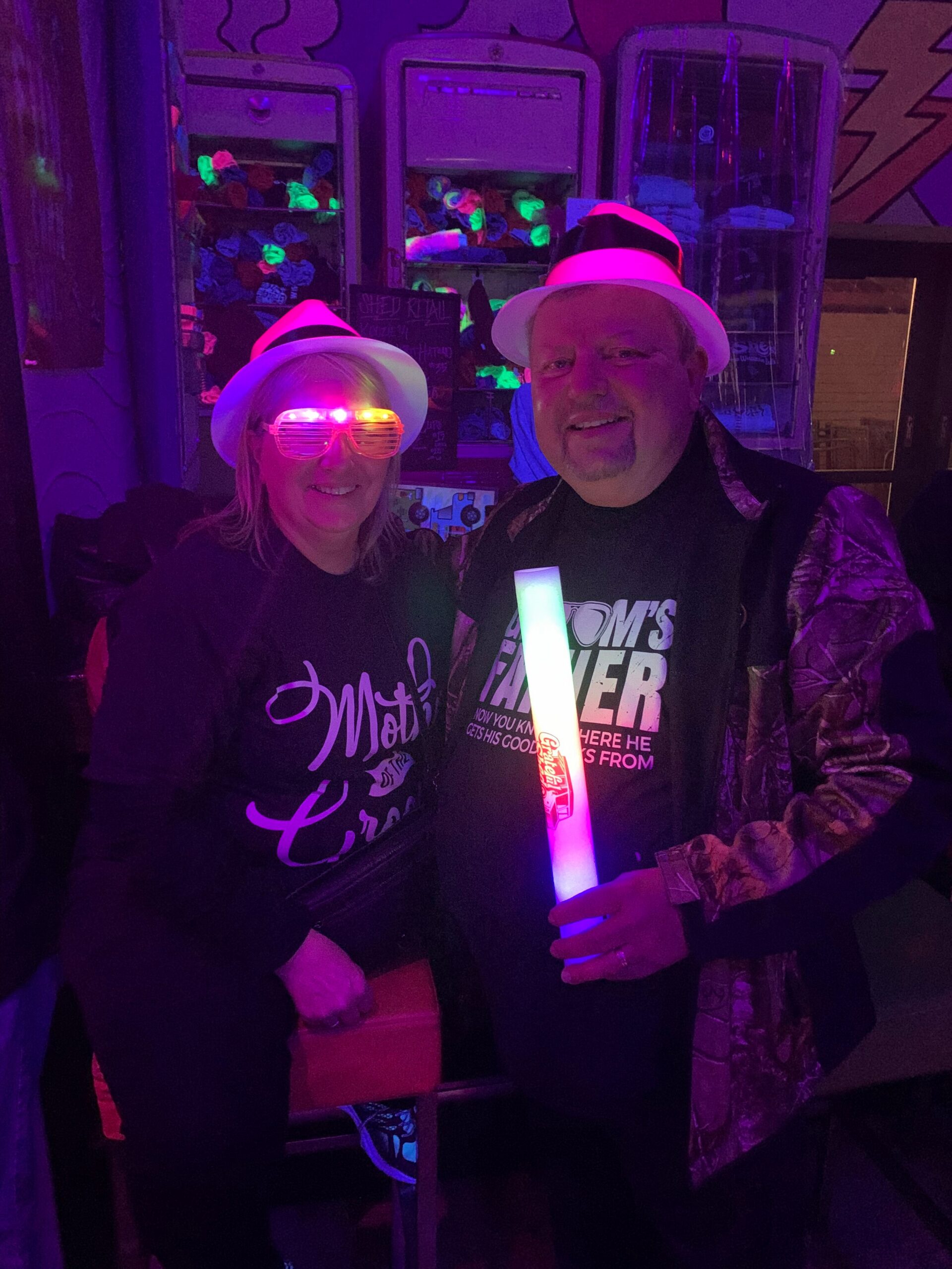 Two smiling adults in a neon-lit setting, wearing white fedoras and glow accessories; the woman wears pink glow glasses and the man holds a glowing tube, with arcade machines in the background.