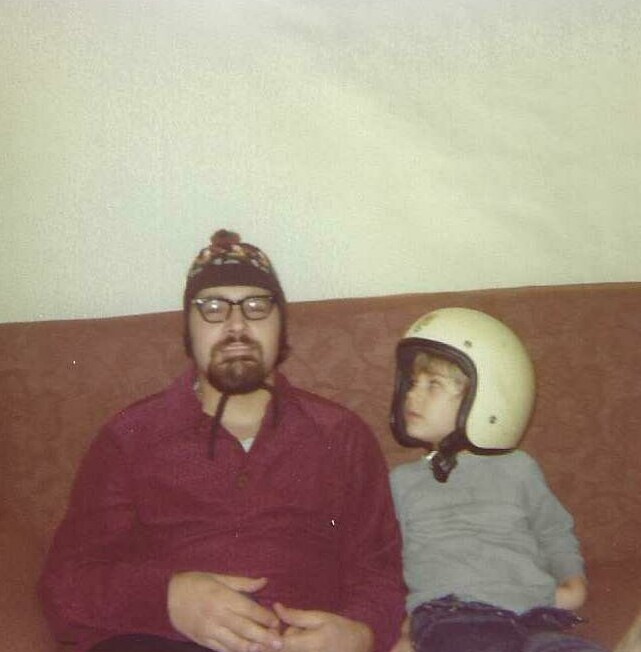 Man with glasses and a knit hat sitting on a couch next to a boy wearing a white helmet.