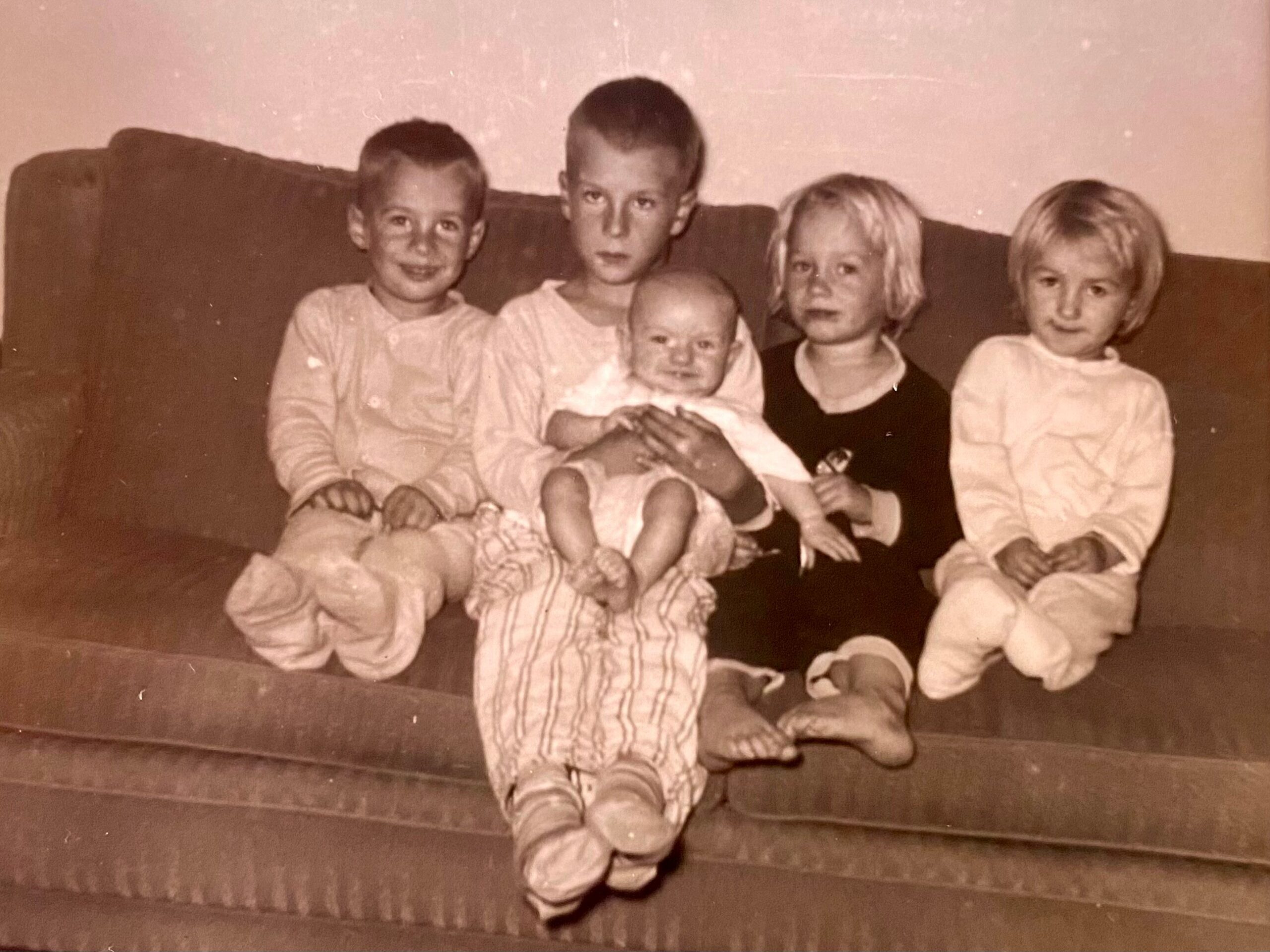 Vintage photo of five children sitting closely on a couch, with a baby in the center and two older kids on each side of him/her.