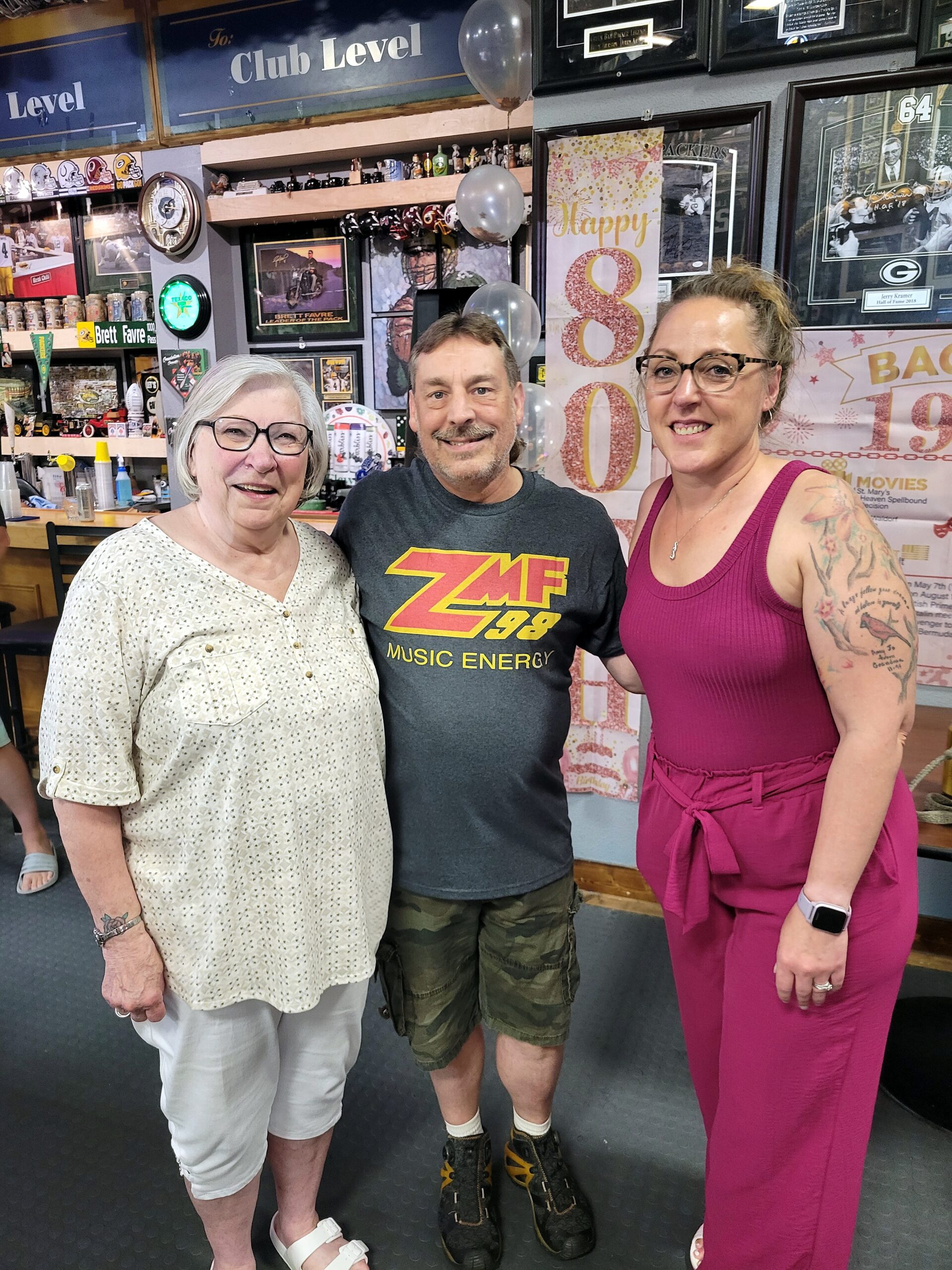 Three adults smiling for a photo in a memorabilia-filled room with framed sports photos, balloons, and a large pink '80' banner in the background.