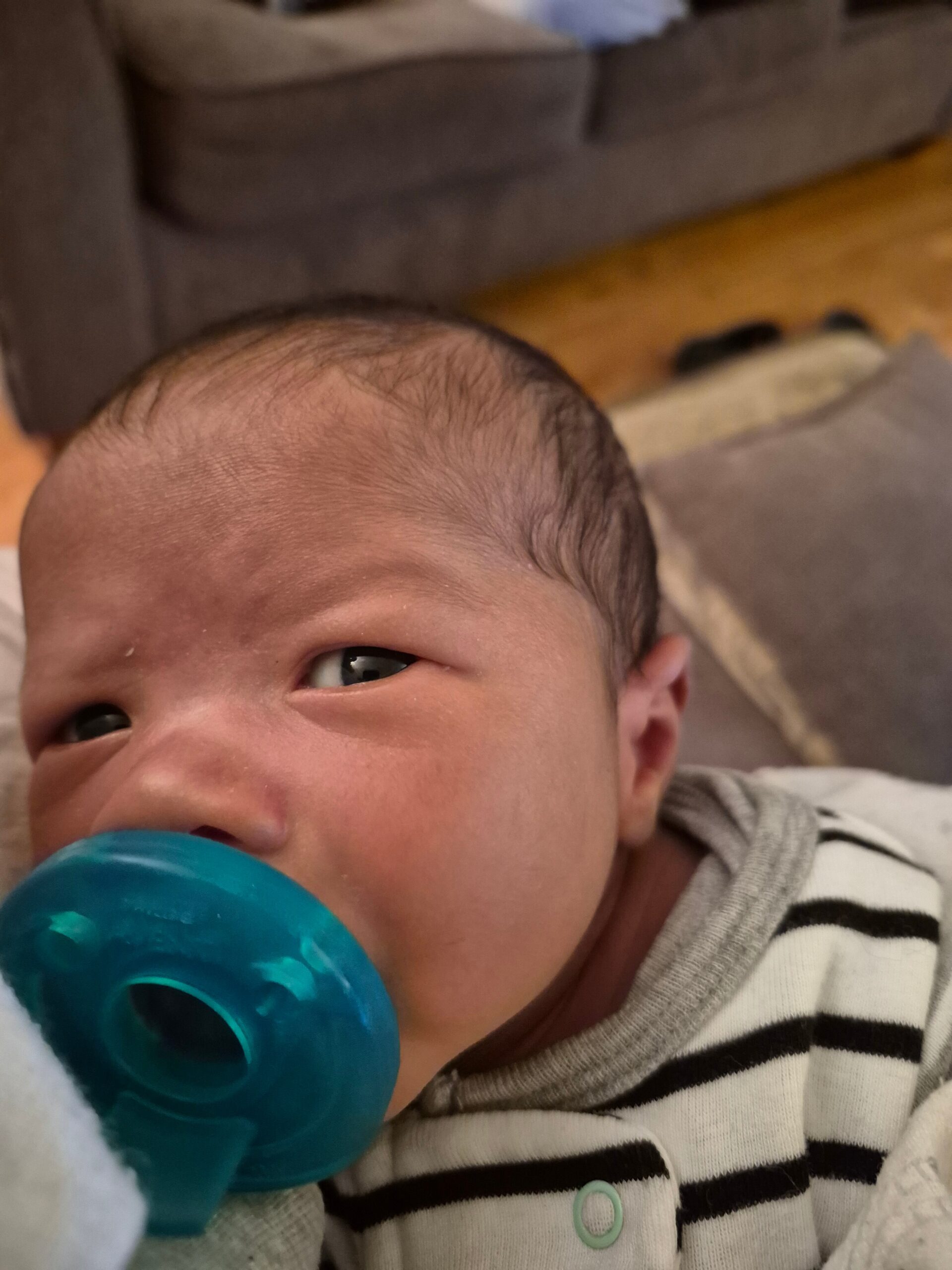 Close-up of a baby with a blue pacifier in the mouth, wearing a striped onesie on a soft surface.