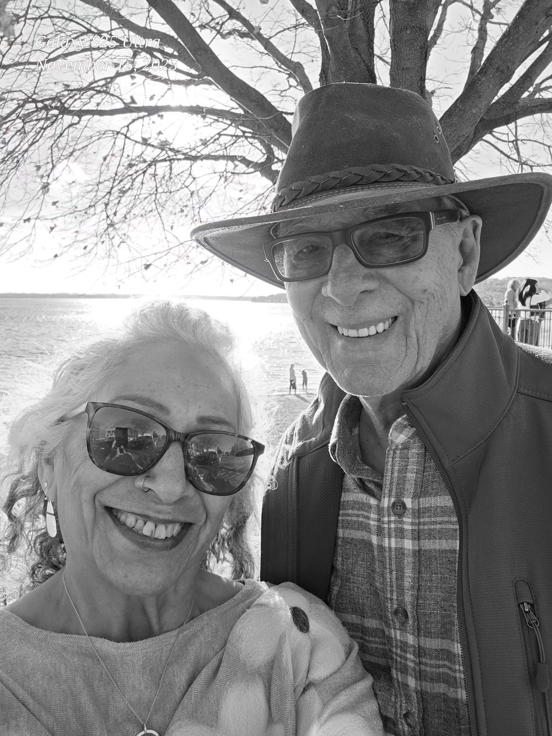 Two smiling older adults take a close-up selfie outdoors, wearing sunglasses with a tree and water in the background; an overlay reads 'Galaxy S25 Ultra November 15, 2025'.