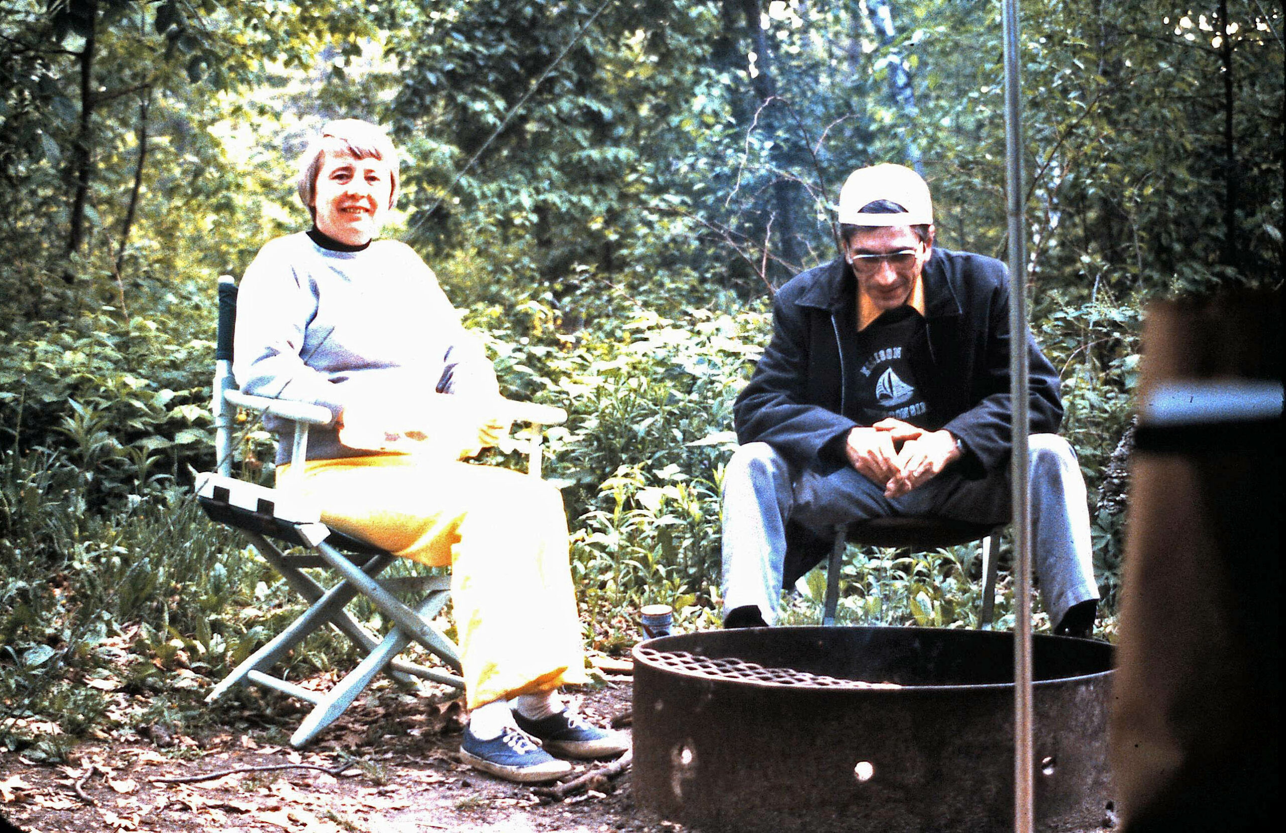 Two people sit in folding chairs in a wooded outdoor area, near a round metal fire pit, smiling at the camera.