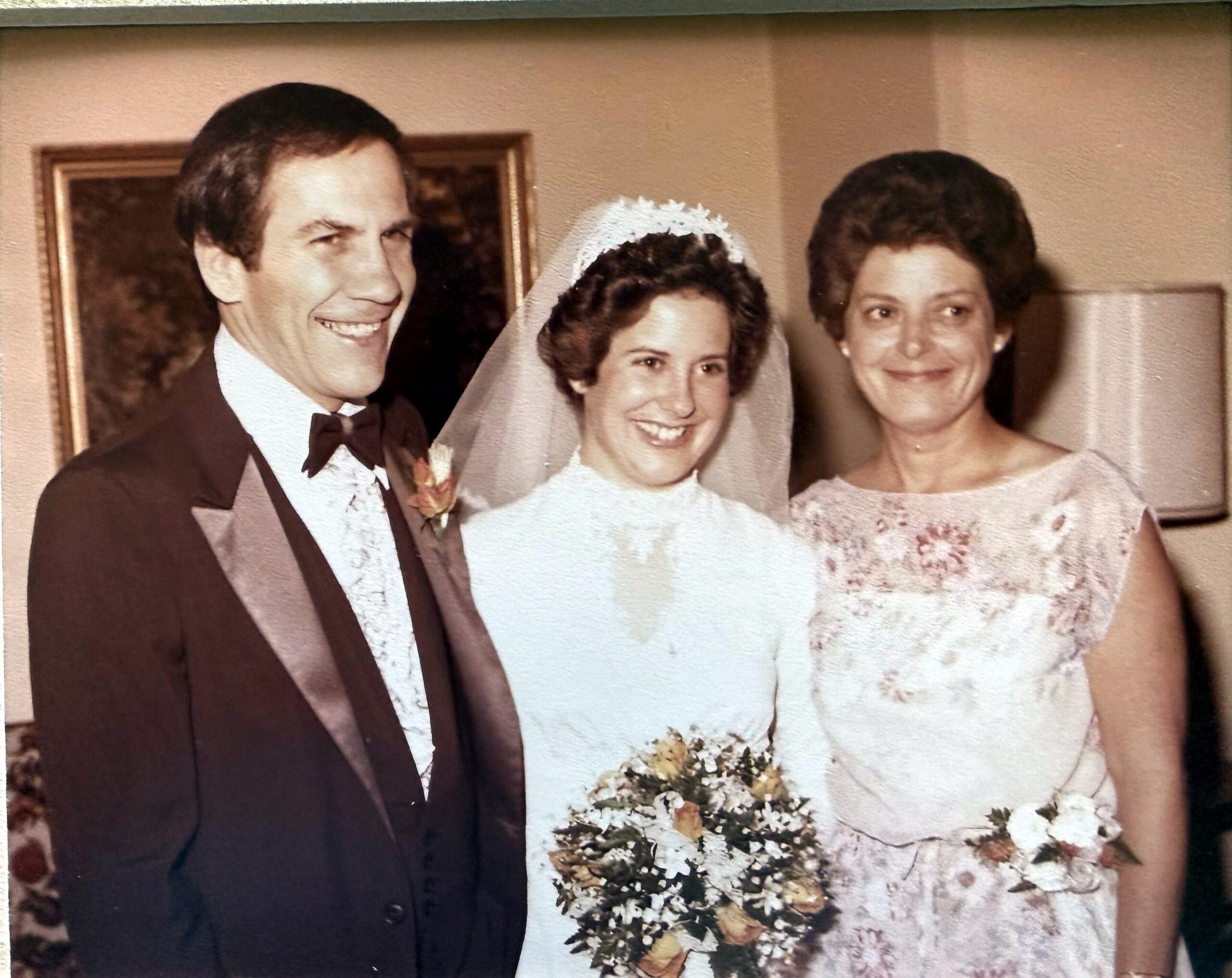 Three people at a wedding: a smiling groom in a tux, a bride in a white dress and veil, and a bridesmaid in a floral dress holding a bouquet.