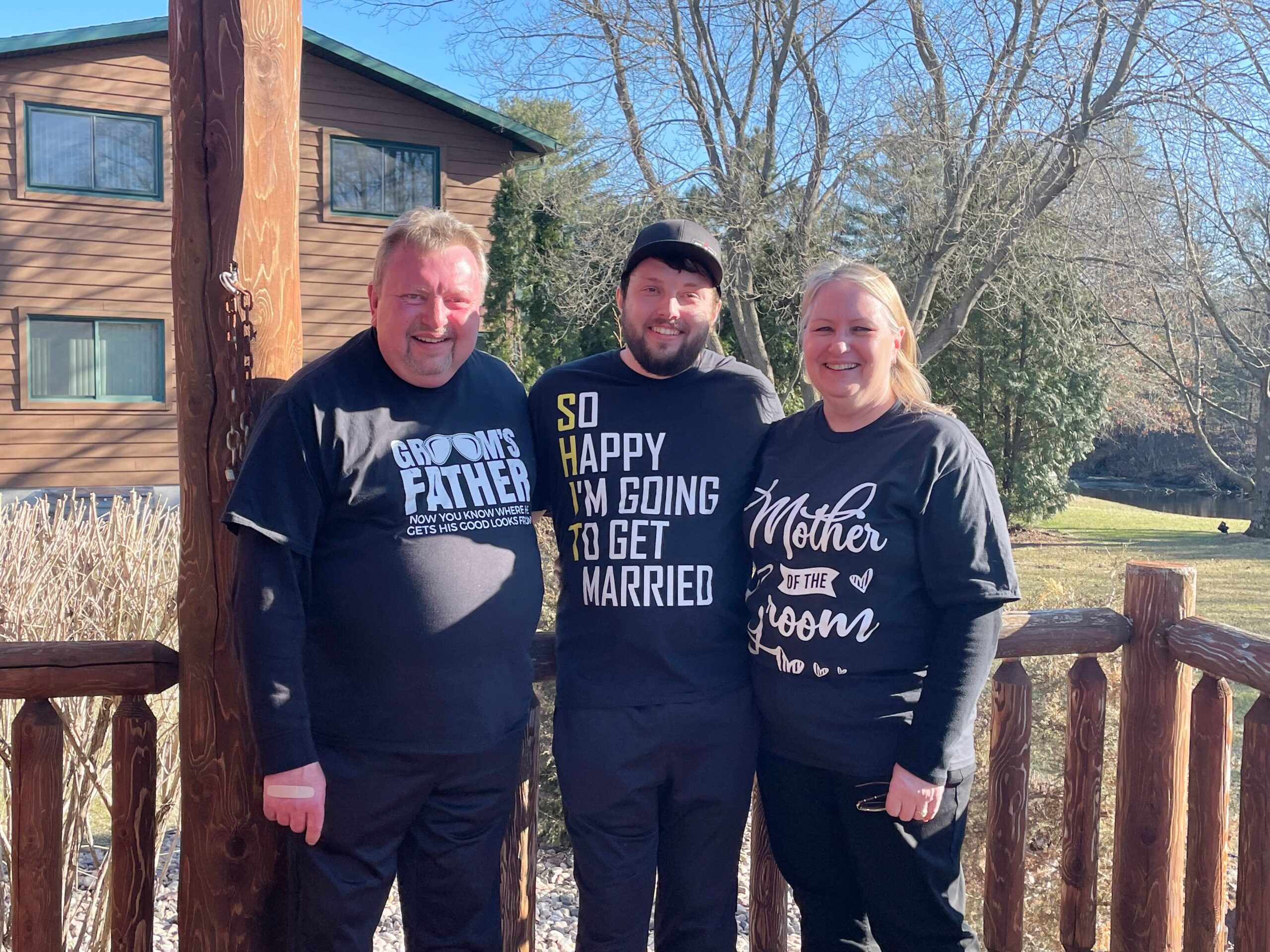 Three adults posing together on a wooden deck outdoors; left man in a 'Groom's Father' shirt, middle man in a 'So Happy I’m Going to Get Married' shirt, right woman in a 'Mother of the Groom' shirt.