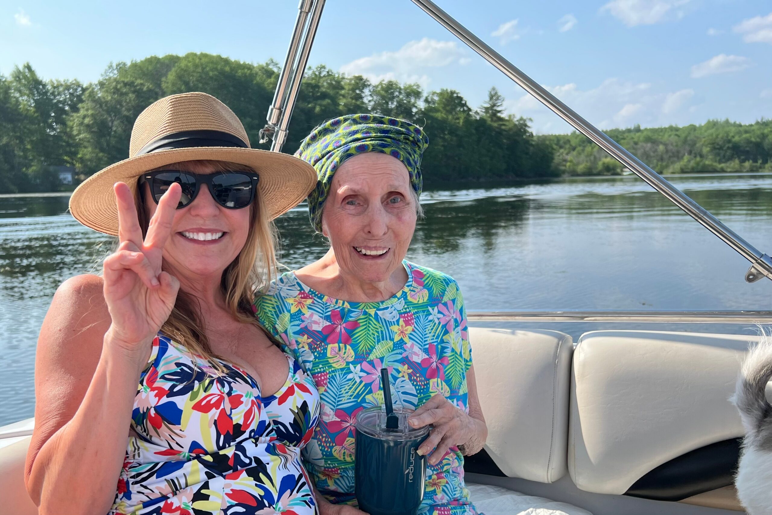 Two women on a boat smiling for a selfie; one wears a straw hat and sunglasses and makes a peace sign, the other has a colorful head wrap and holds a drink with a straw in bright sunlight.