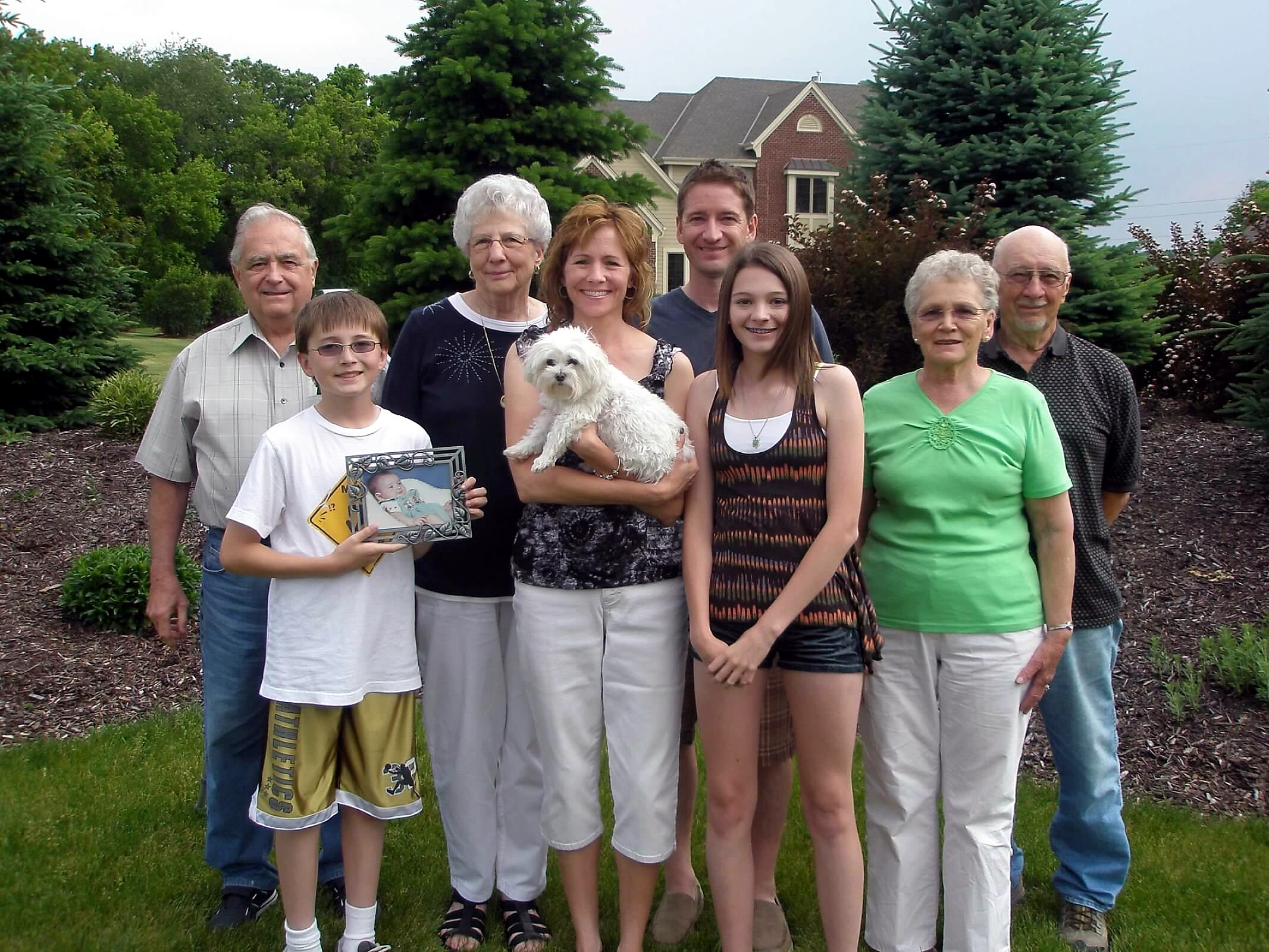 Group of eight people standing together outside in a yard, a woman in the center holding a small white dog, with trees and a house in the background.