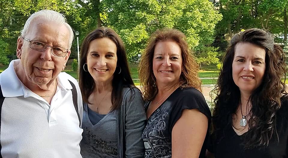 Four adults smiling together outdoors in a sunny park with green trees in the background.