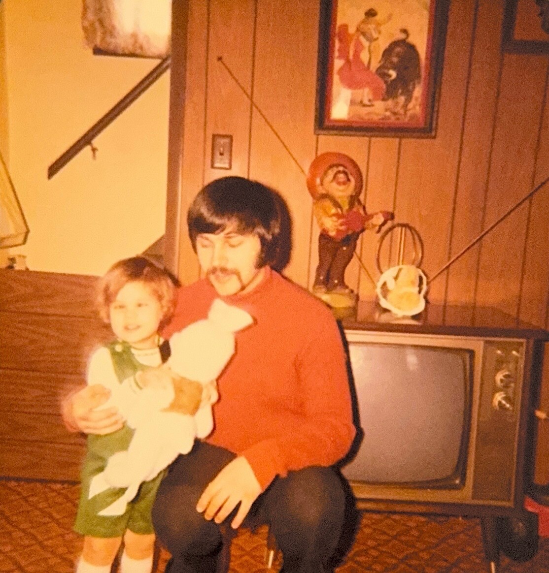 Man kneeling beside a young child in a retro living room with wood-paneled walls and a vintage TV in the background, posing for a family photo.