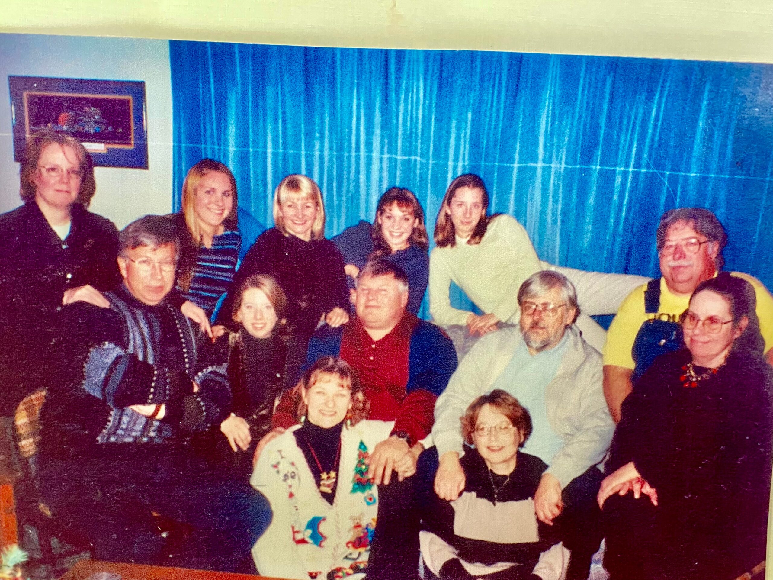 Group of people posing for a photo in a room with blue curtains in the background, smiling faces gathered together on a couch and floor.