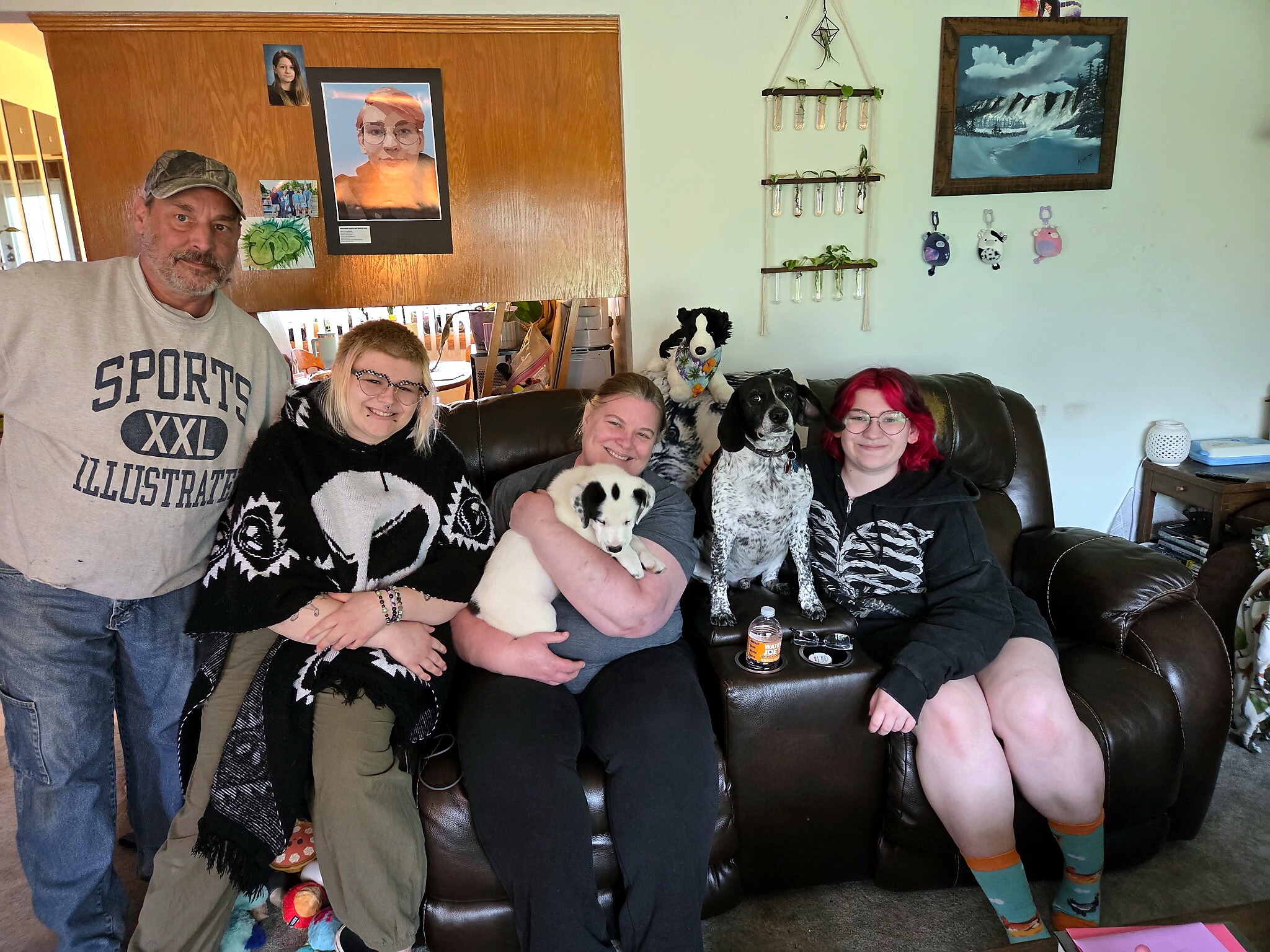Five people sit on a brown leather couch indoors, smiling with two dogs on their laps in a cozy living room.
