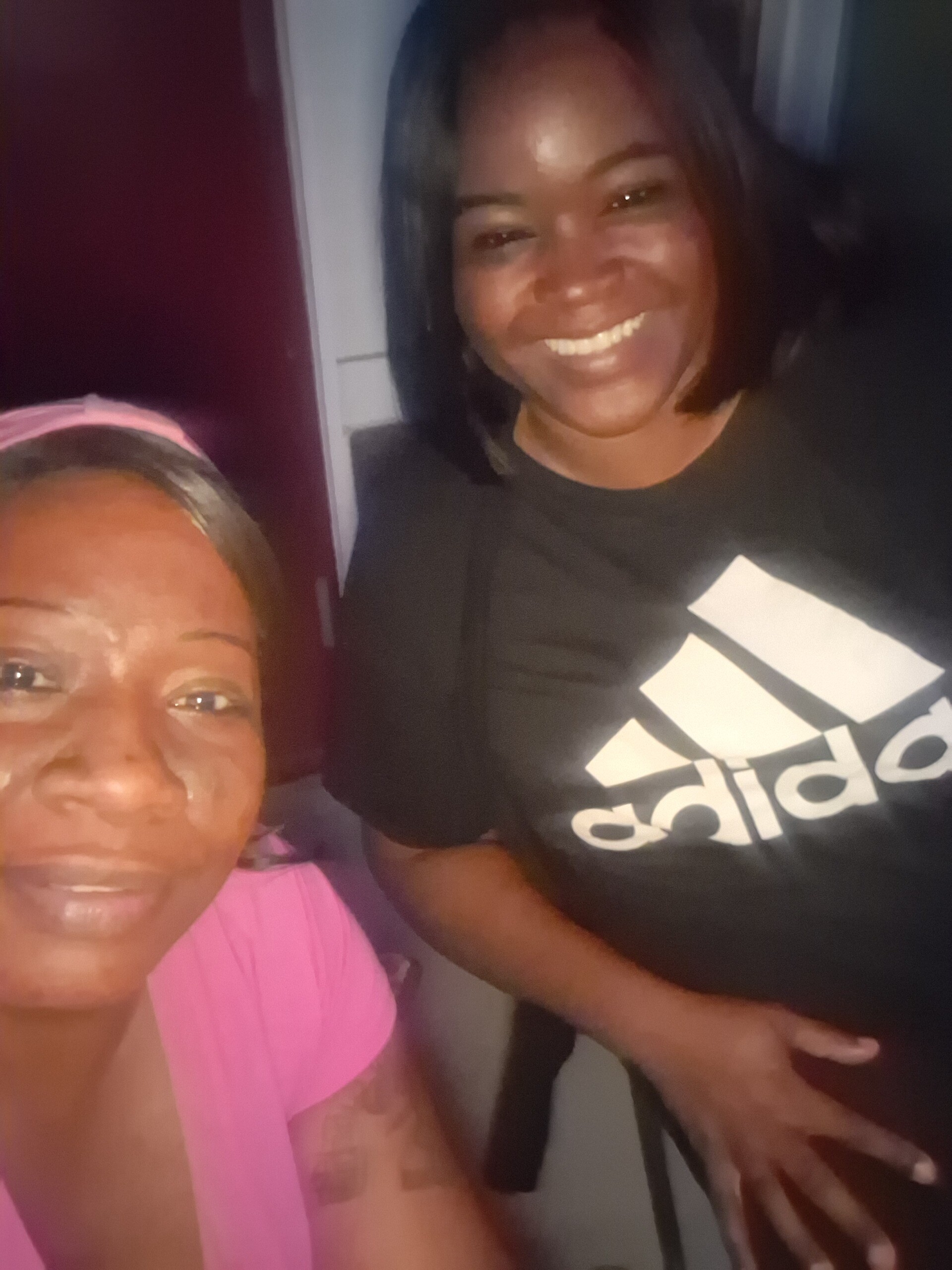 Two smiling people posing for a selfie, one in a pink top on the left and the other in a black Adidas shirt on the right.