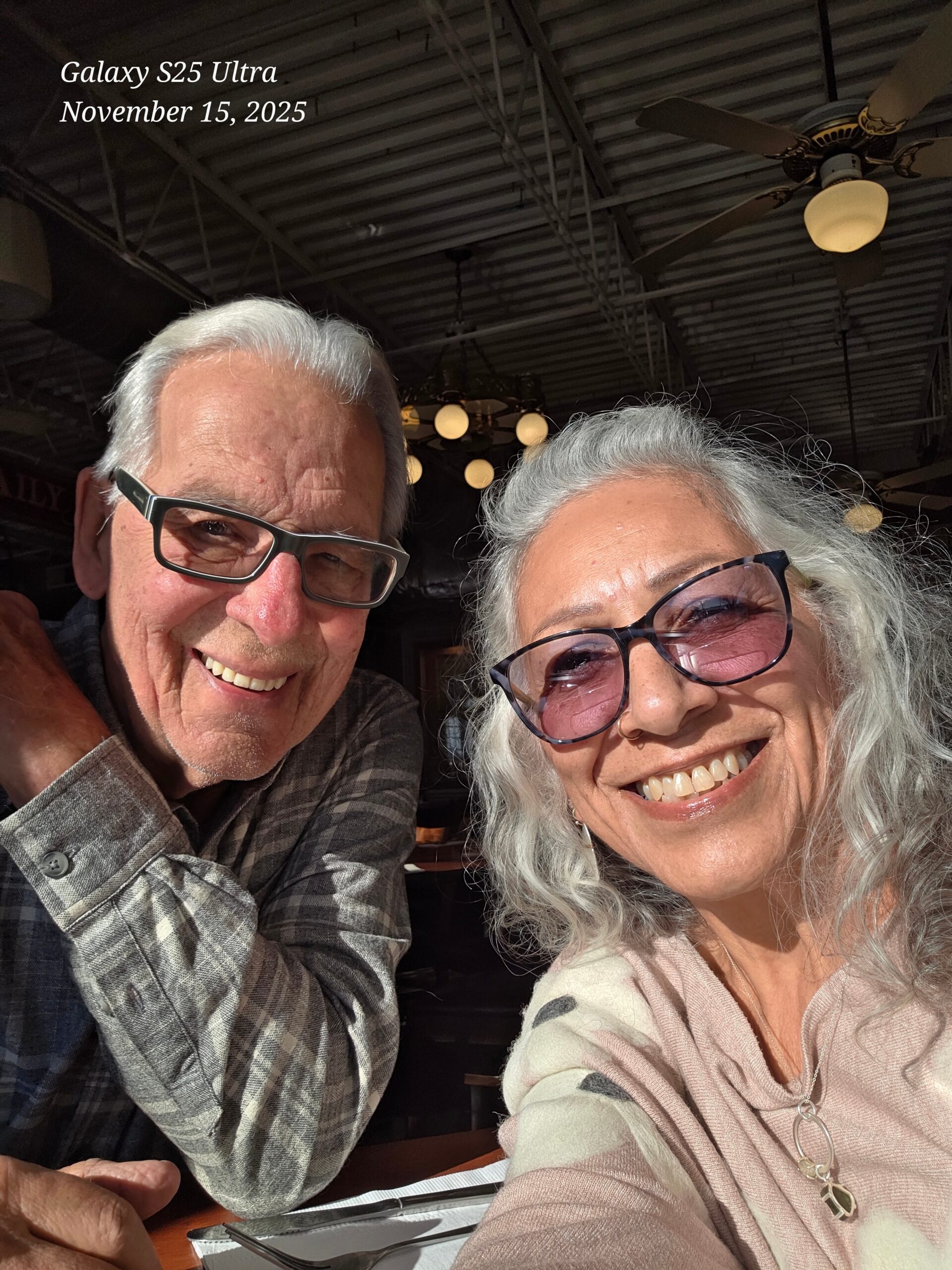 Smiling elderly couple taking a selfie at a restaurant with ceiling fans in the background.
