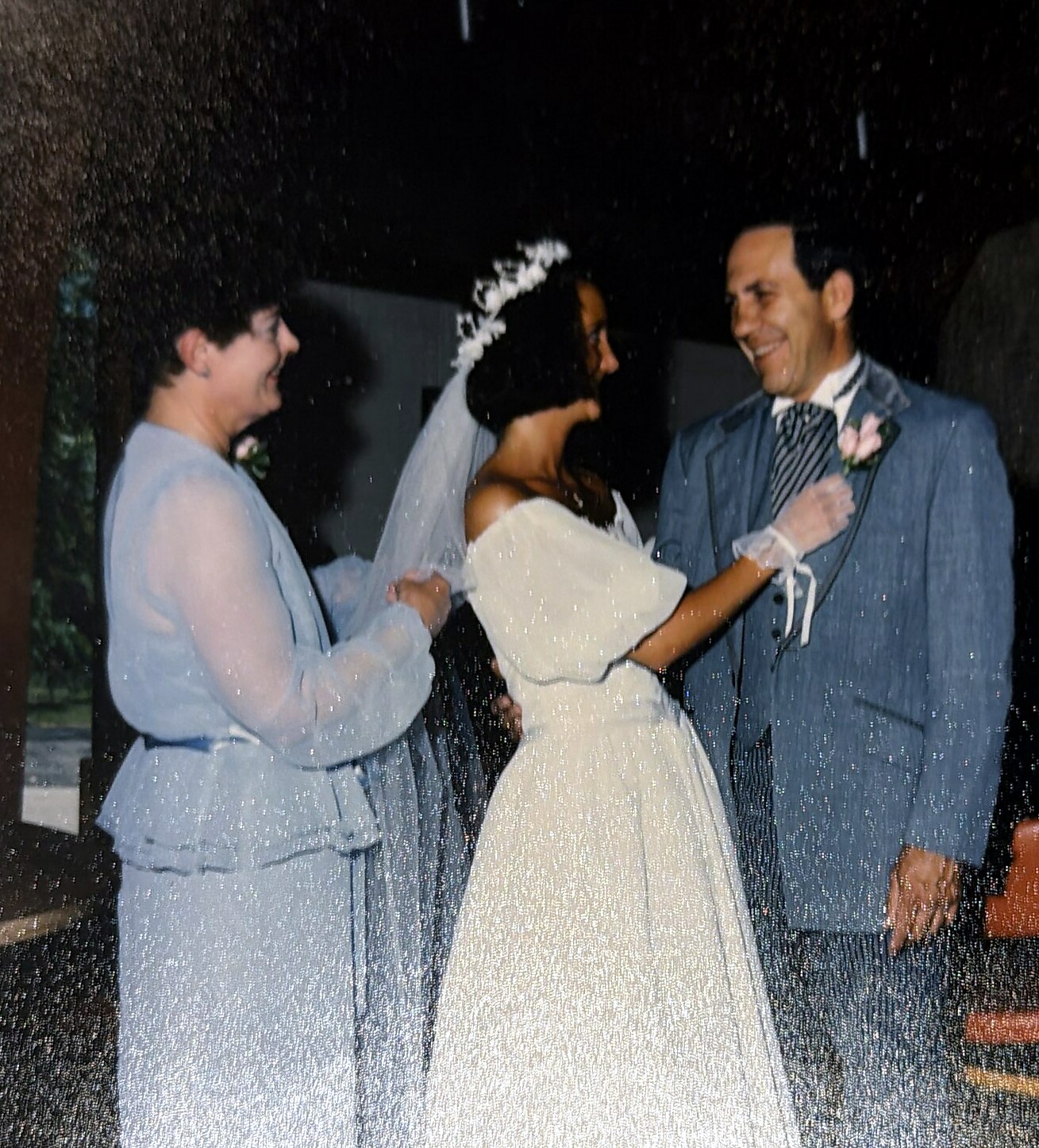 Bride in a white wedding gown and veil shares a moment with a smiling groom in a blue suit during a nighttime wedding, flanked by two attendants in pale blue outfits helping with the veil.