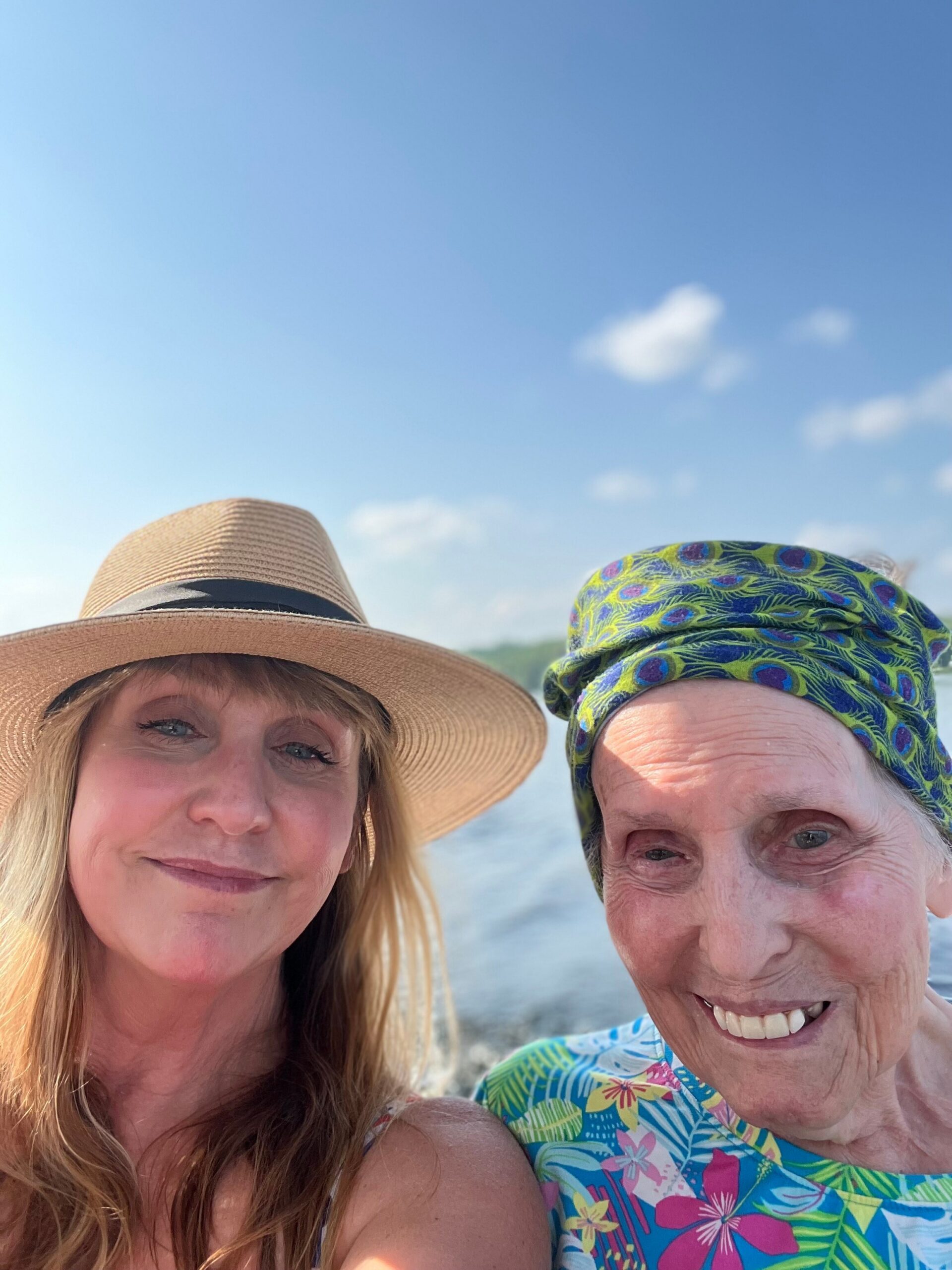 Two women smiling at the camera on a sunny day at the beach, one wearing a wide-brim straw hat and the other a colorful patterned headscarf.