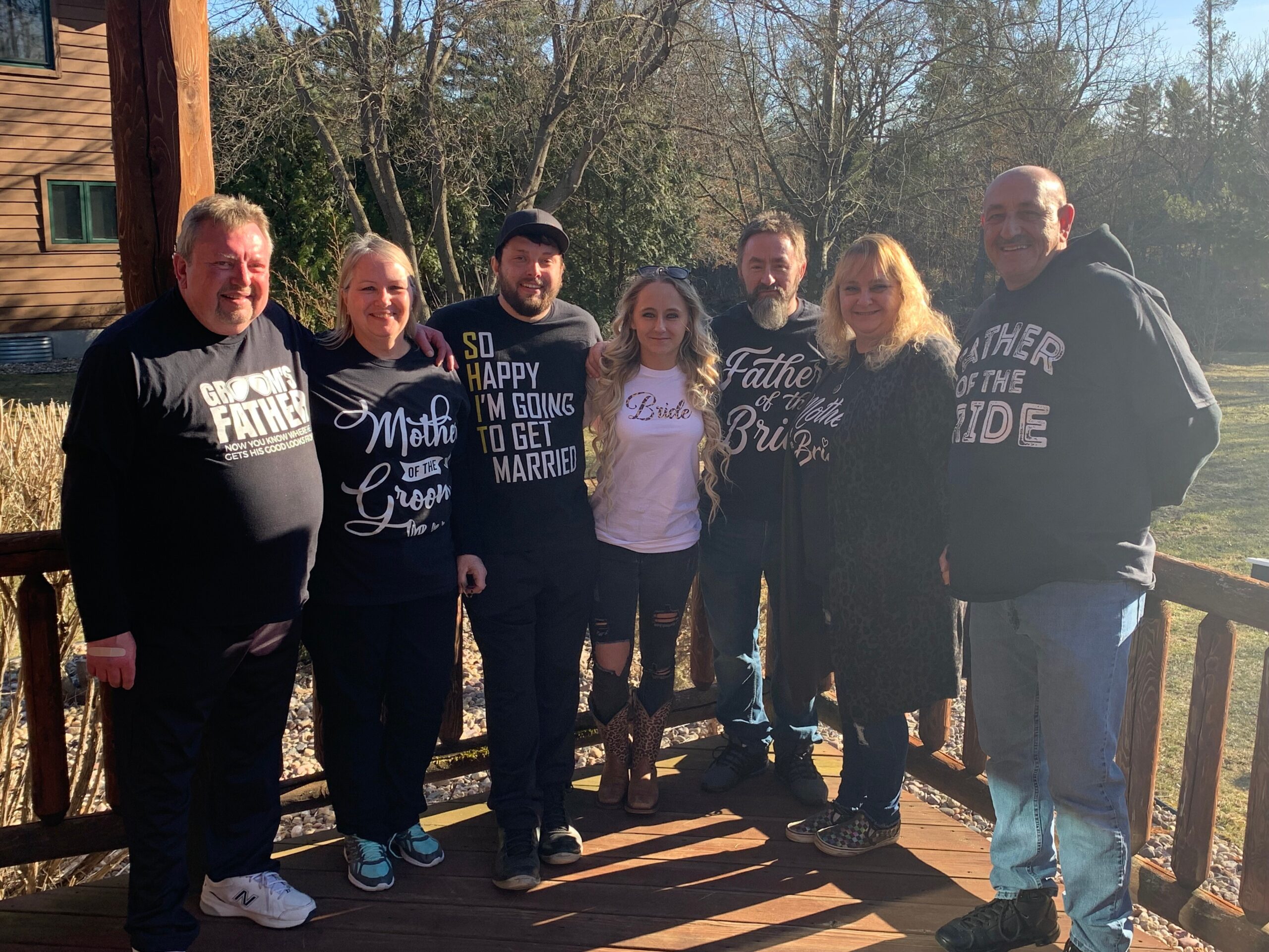 Seven adults stand arm-in-arm on a wooden deck outdoors, wearing casual black shirts with wedding-themed text, smiling with a wooded yard and house in the background.